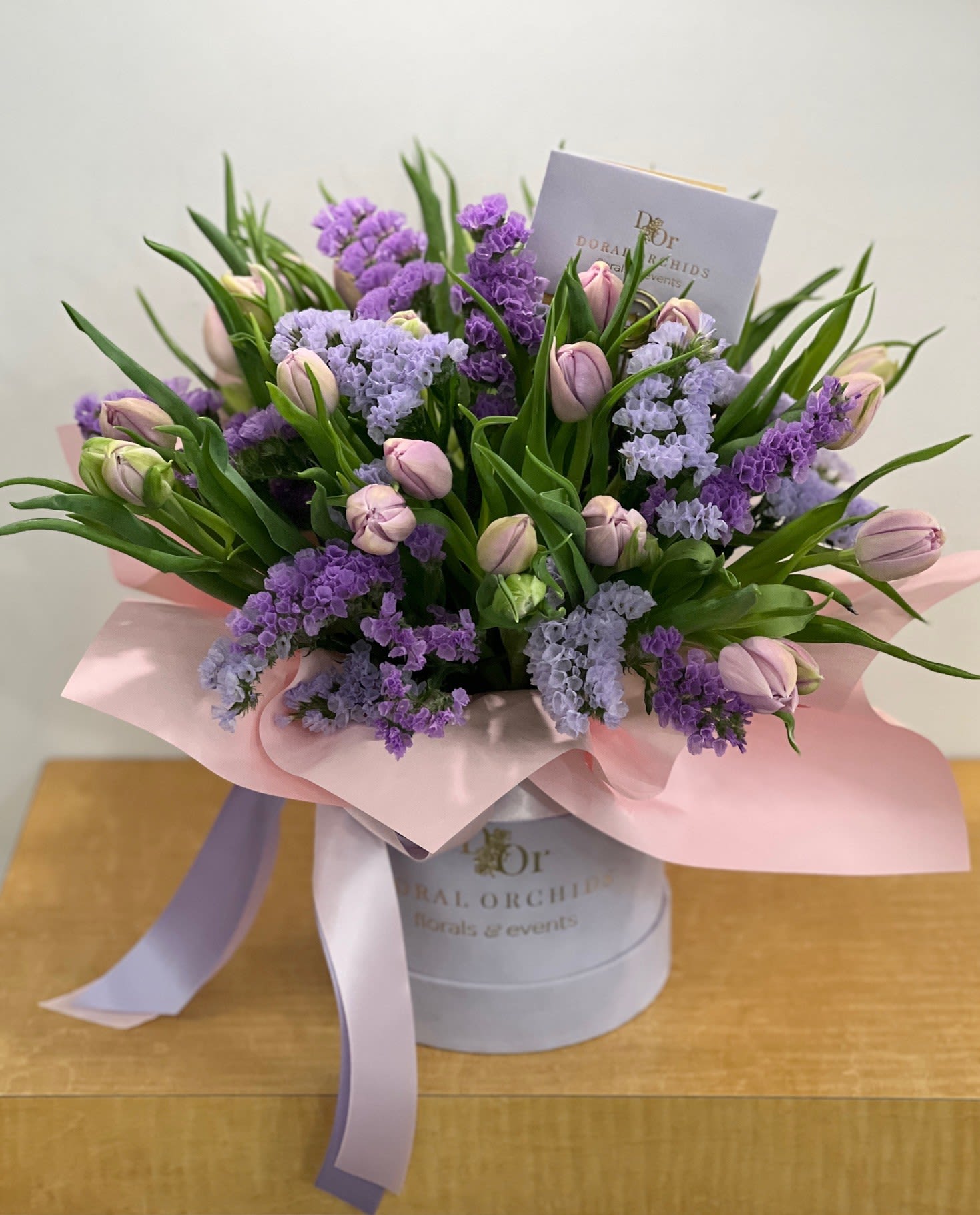 Tulips in a box - Lavender Dreams ™ - A soft pastel arrangement of lilac tulips, lavender blooms, and fresh greenery, gathered in a round hatbox with satin ribbon. It’s a refreshing design full of springtime charm.