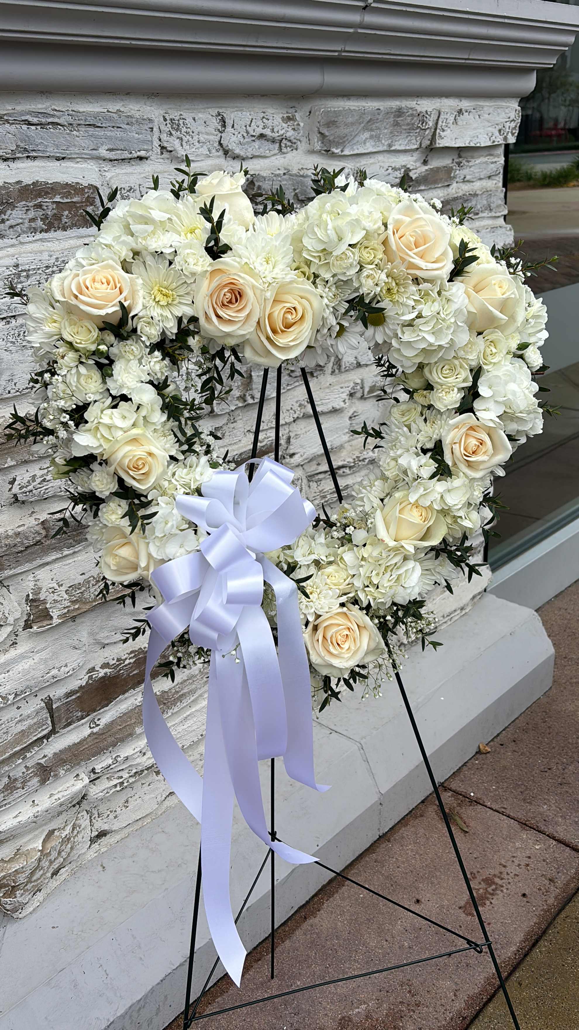 Heart-shaped white floral wreath with cream roses and a lavender ribbon on a stand