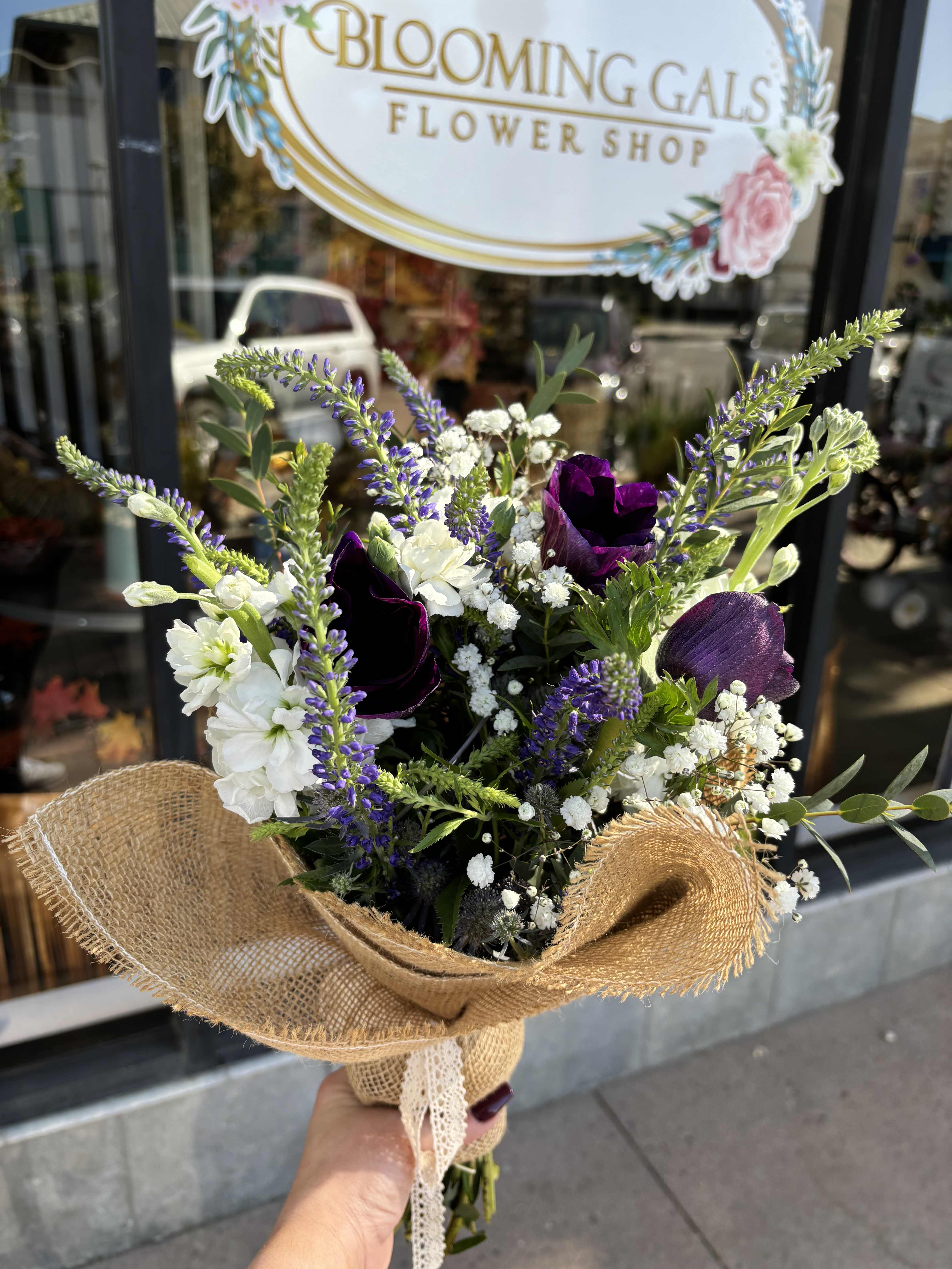 Handheld bouquet of purple, white, and green flowers wrapped in burlap
