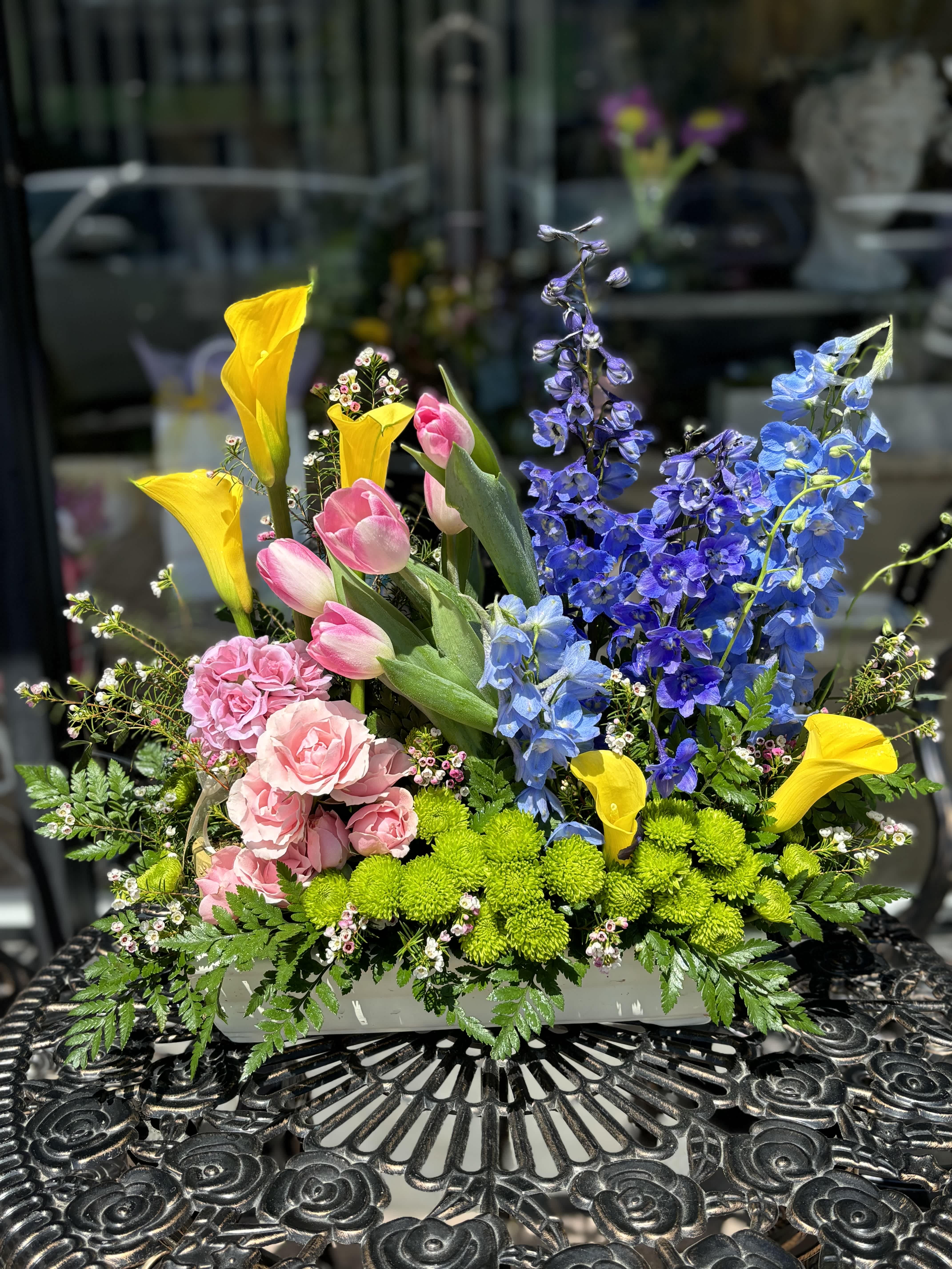 Colorful mixed flower arrangement with tulips, calla lilies, and blue delphinium in a white container