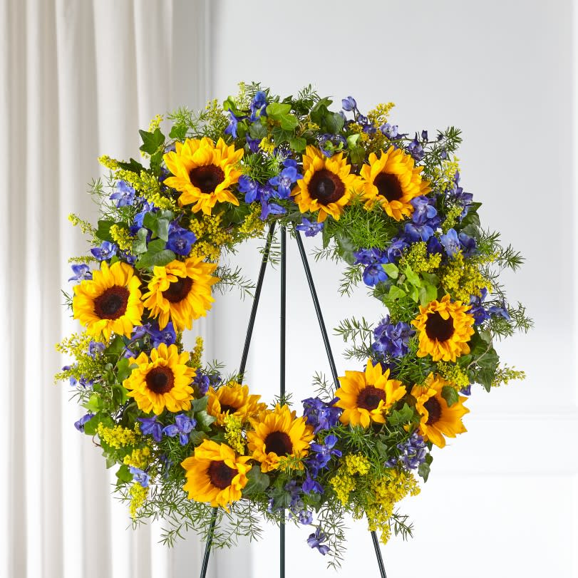 The FTD® Bright Rays™ Wreath  - The beautiful sunflowers in our Bright Rays™ Wreath offers a display of admiration, loyalty and hope. Each bright bloom is complemented by lush greens and blue delphinium blooms. Their uplifting nature reminds loved ones of your support and the brighter days ahead as they navigate through a difficult time.  Details: o Wreath is approximately 26" in diameter 