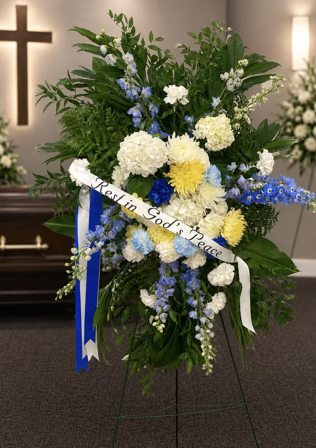 Standing sympathy spray with white, blue, and yellow flowers and a "Rest in God's Peace" ribbon