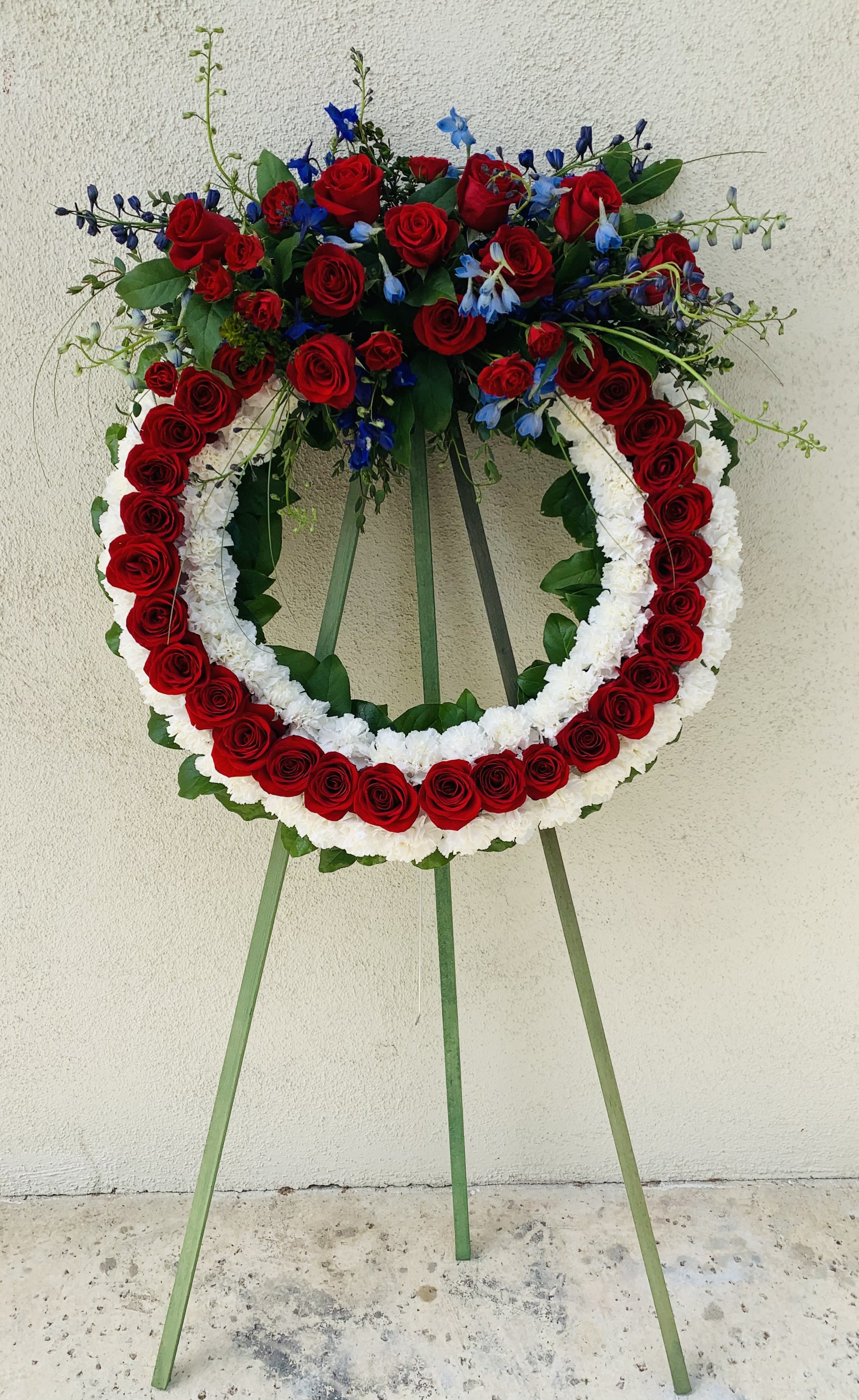 Standing floral wreath of red roses, white flowers, and blue accents on an easel