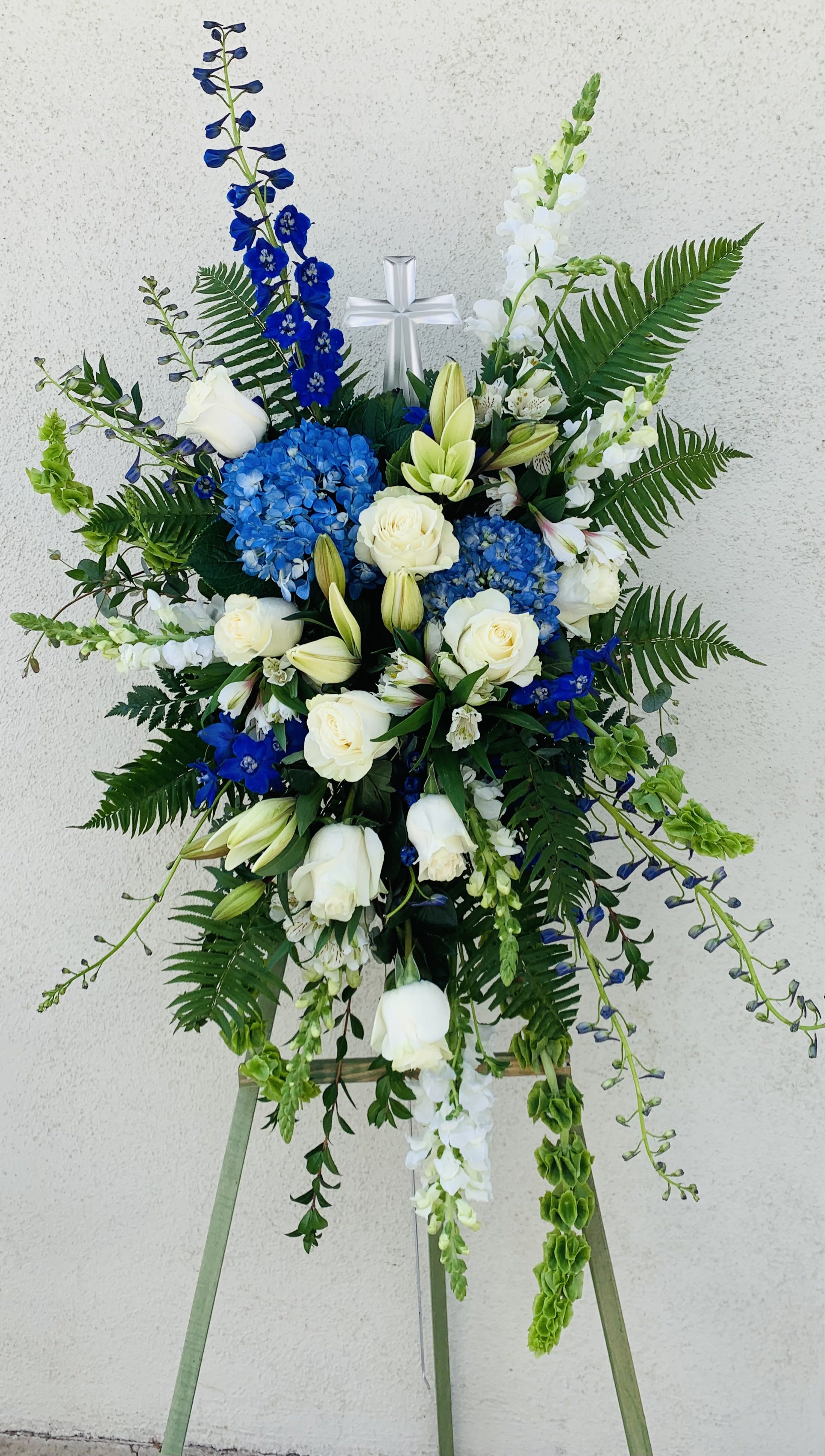 Tall floral standing spray with white roses, blue hydrangeas, and a silver cross