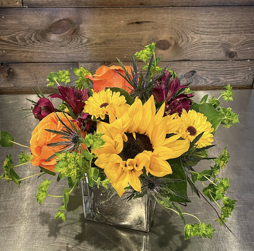 State Fair - A kaleidoscope of color with fun bright tones, including sunflowers, roses, alstromeria, viking poms and greeney in a small square vase. Perfect to be placed on a desktop for any occasion. Approx. 8" Tall x 10" Wide  