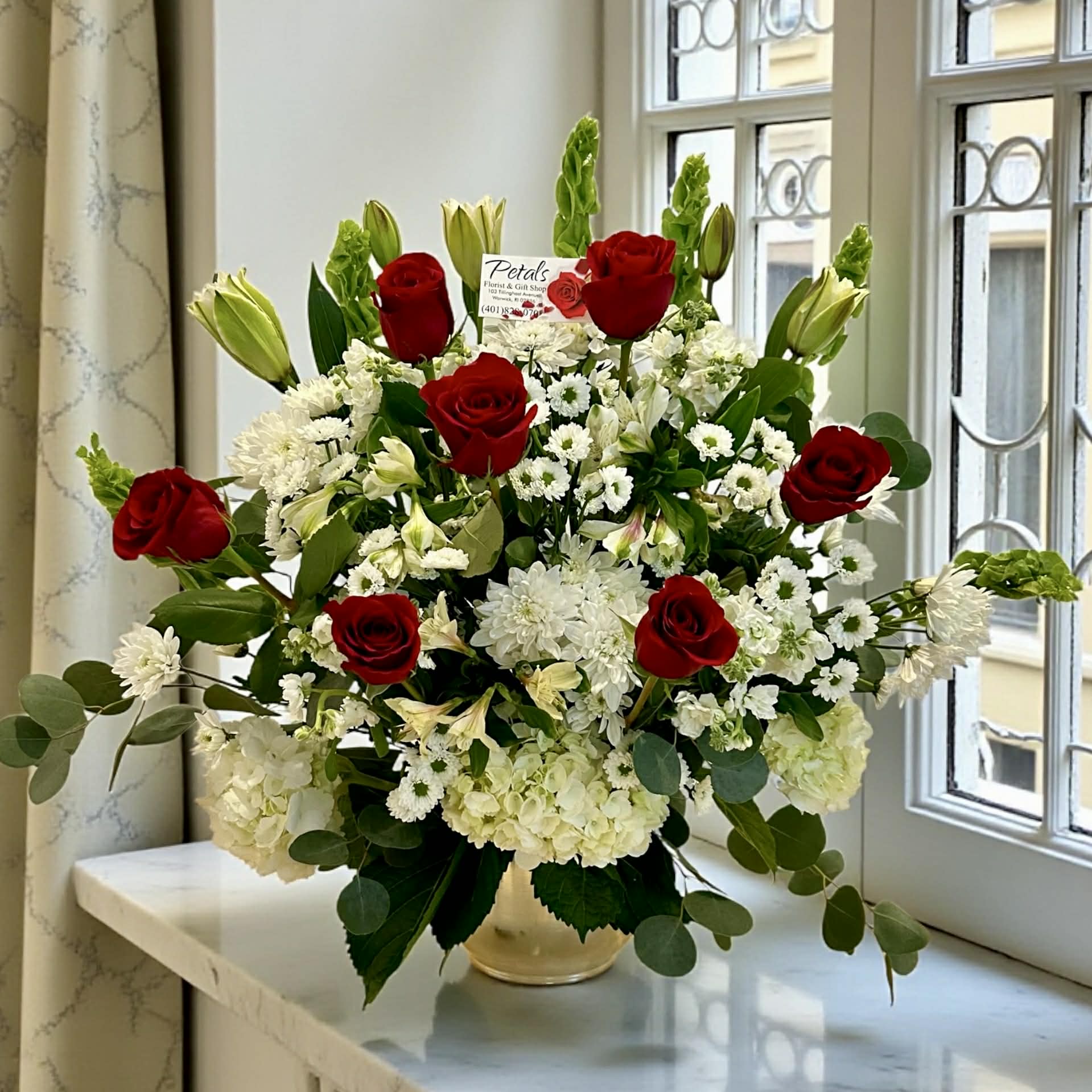 Loving Red Embrace Tribute by Petals - Red roses create a powerful and meaningful tribute, symbolizing courage, respect, and a love that endures beyond loss. Paired with serene white lilies, Bells of Ireland, and abundant white hydrangeas, this elegant, forward-facing arrangement reflects both peace and lasting reverence.  Thoughtfully designed to honor a vibrant life, it offers a heartfelt expression of sympathy and a source of comfort to those who are grieving. While seasonal availability may bring slight variations, the beauty and sentiment of this tribute will always be carefully preserved.
