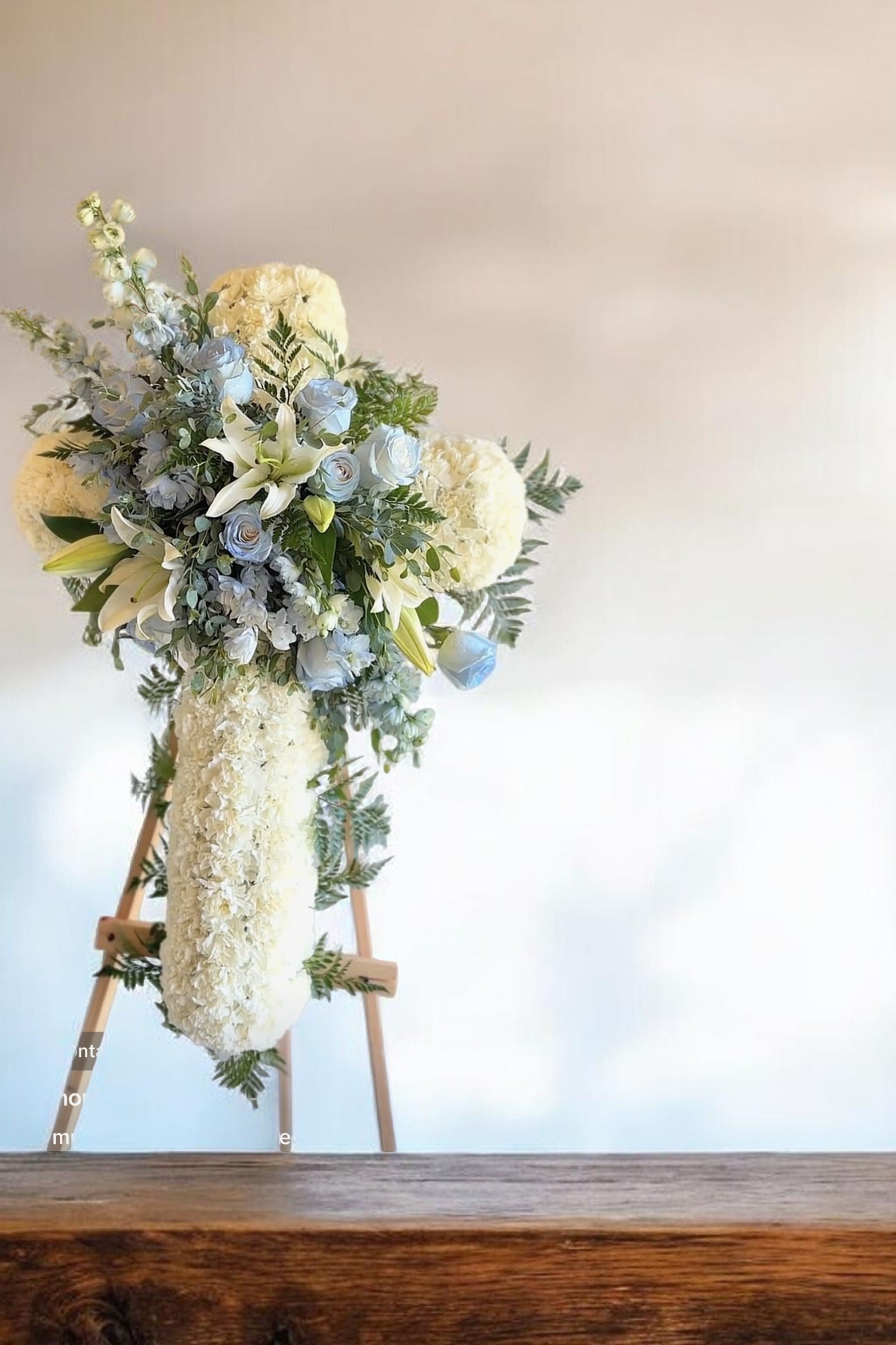 Heavenly Cross Tribute - A beautiful cross-shaped arrangement crafted with soft white blooms and accented with gentle blue roses and lilies. Set on an easel, this elegant design symbolizes faith, peace, and everlasting love—offering a heartfelt expression of sympathy and remembrance.
