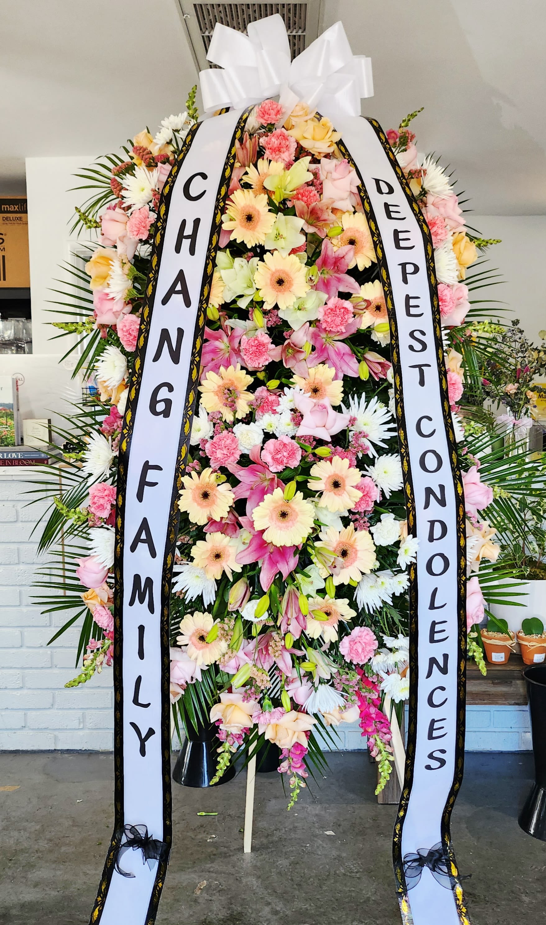 Tall standing spray of pink lilies, carnations, and peach gerberas with condolence ribbons on an easel.