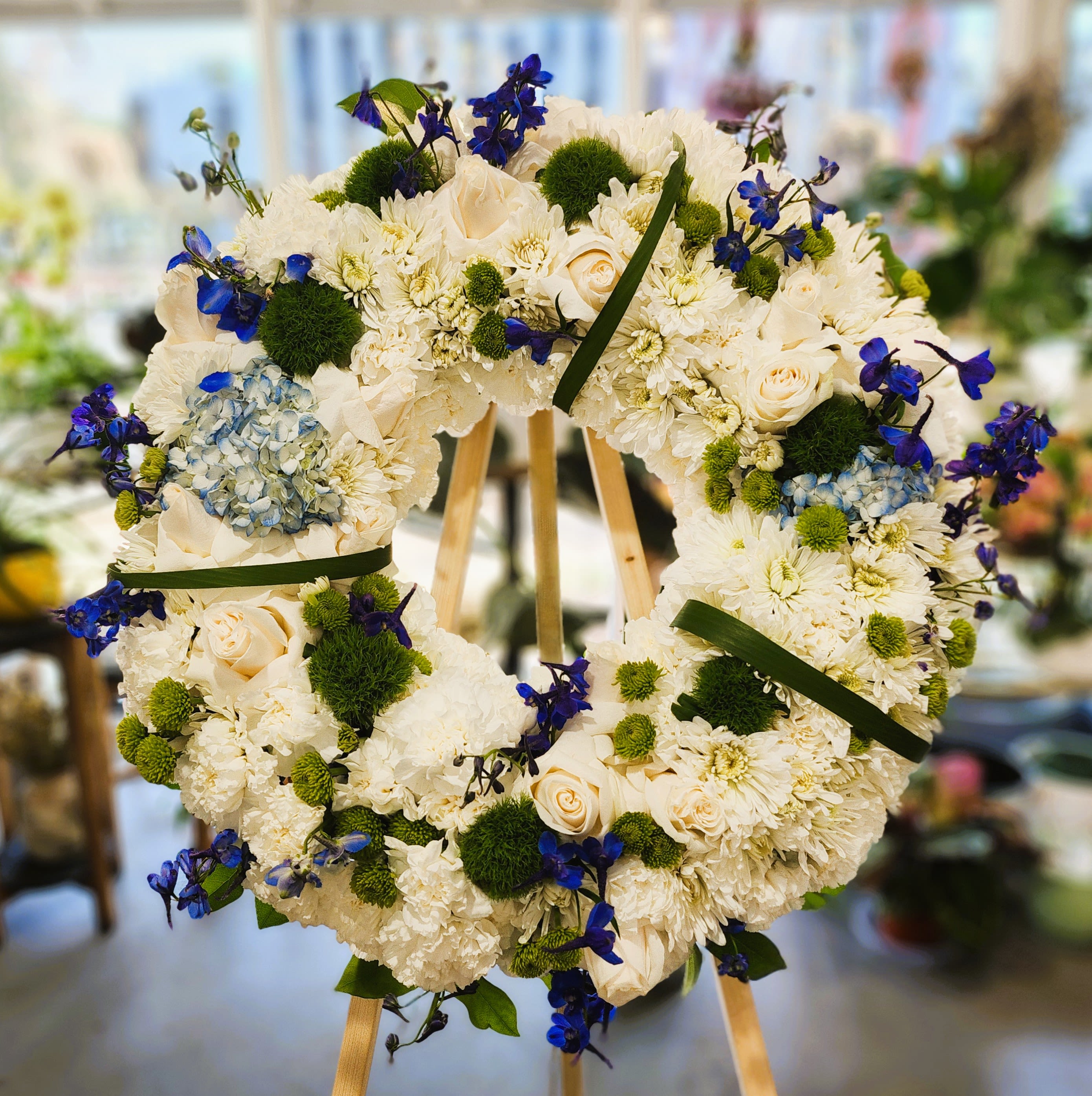 Heart-shaped floral wreath with white blooms, blue accents, and green pom-poms on an easel