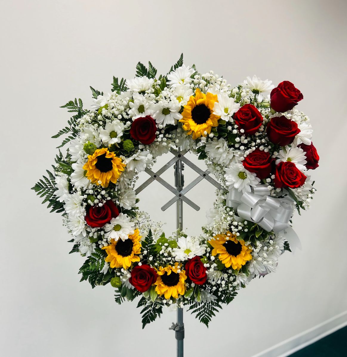 Circular floral wreath with red roses, white daisies, and sunflowers on a stand