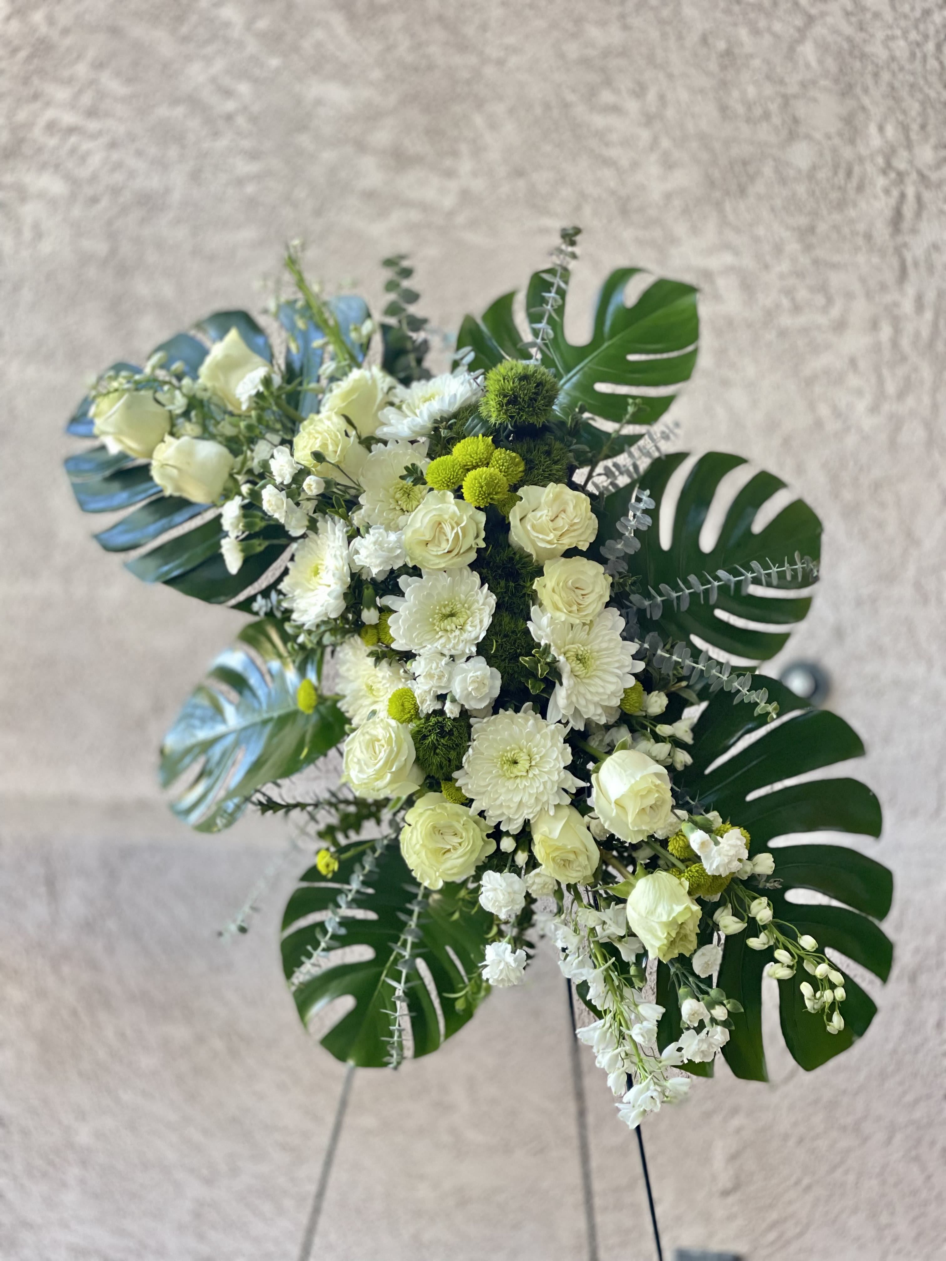 Tall standing spray of white roses and mums with large dark green monstera leaves on an easel