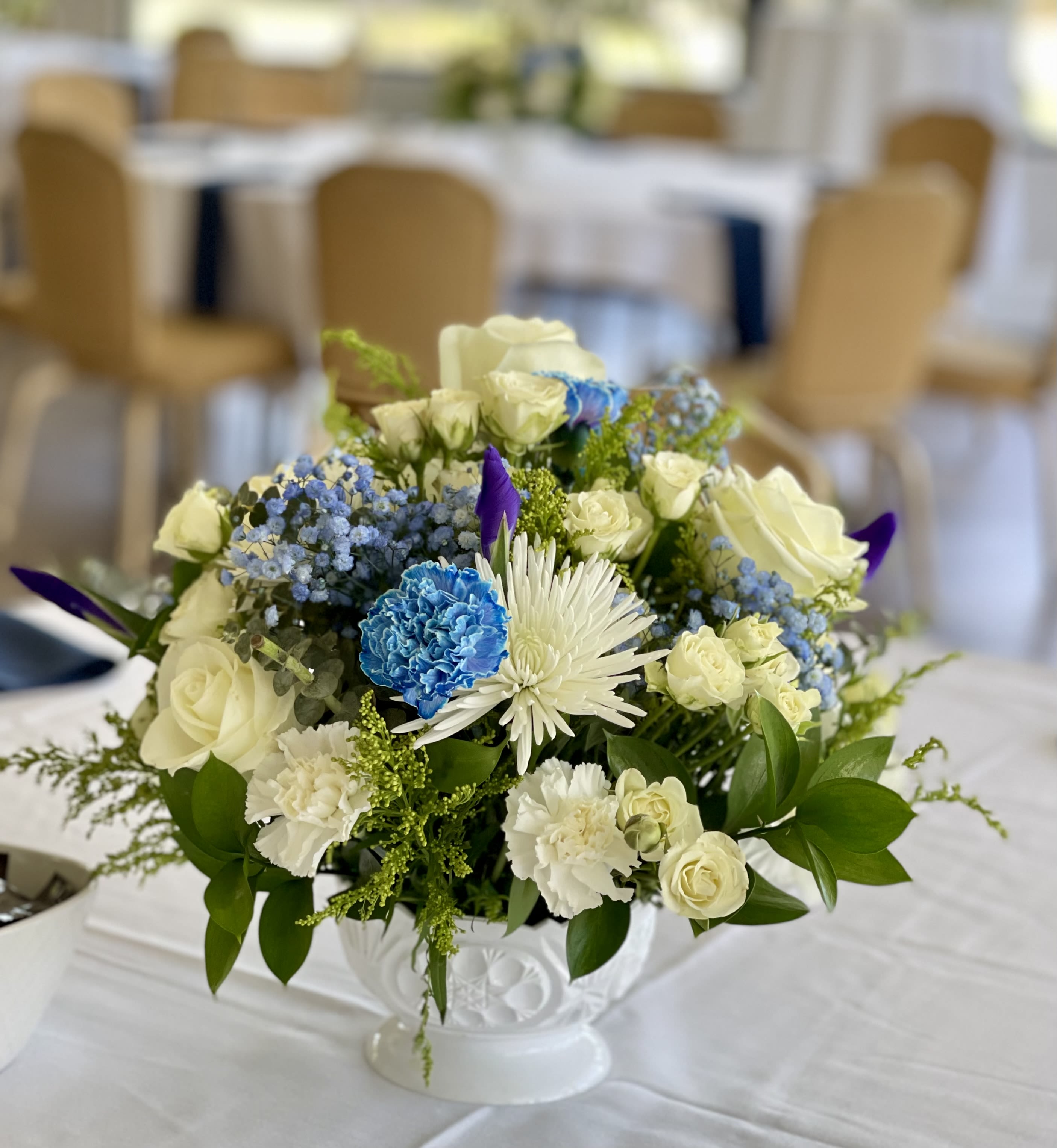 Low centerpiece of white roses, carnations, and blue accents in a white pedestal bowl