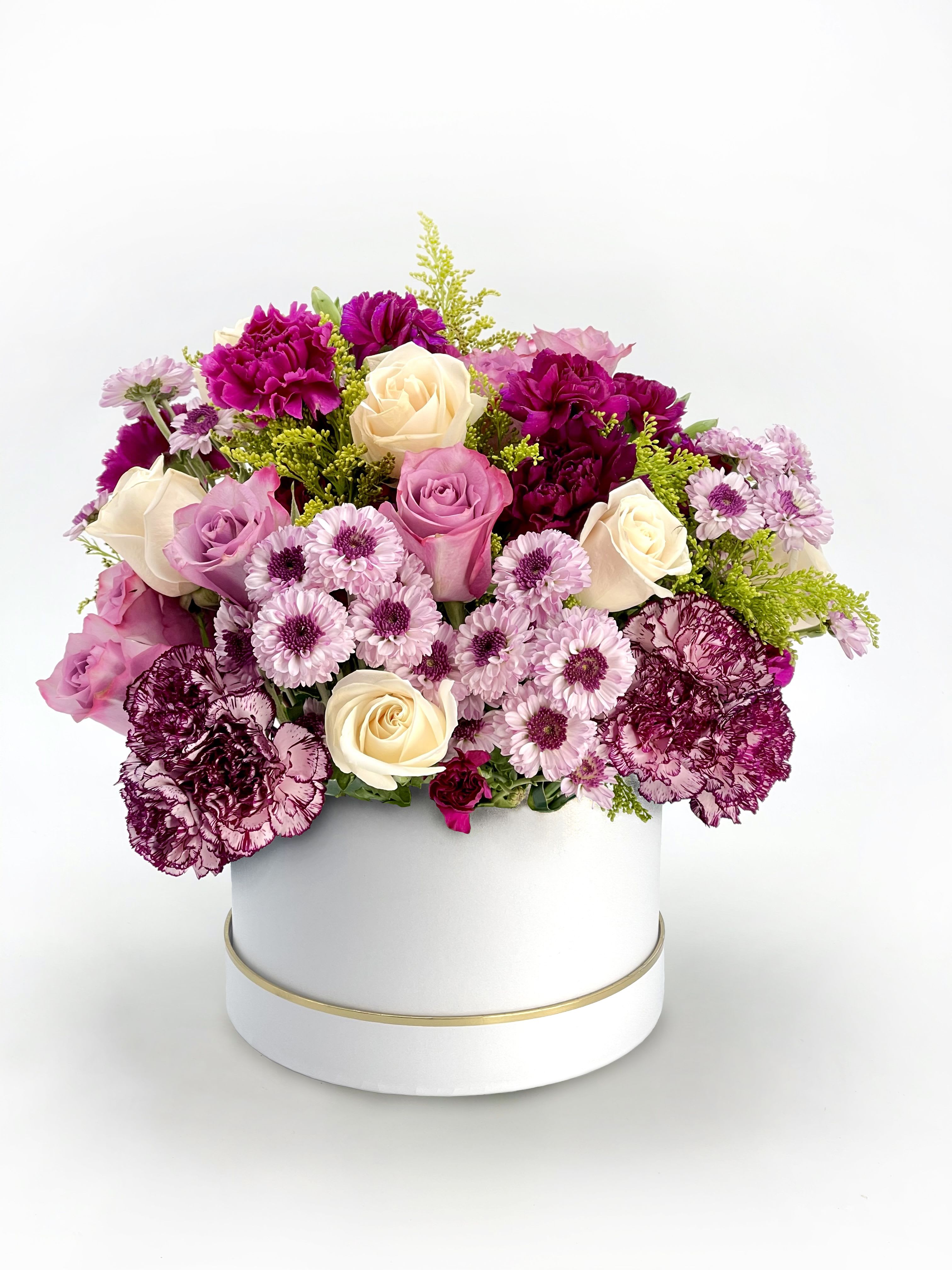 Round arrangement of cream and lavender roses, purple carnations, and mums in a white hatbox container