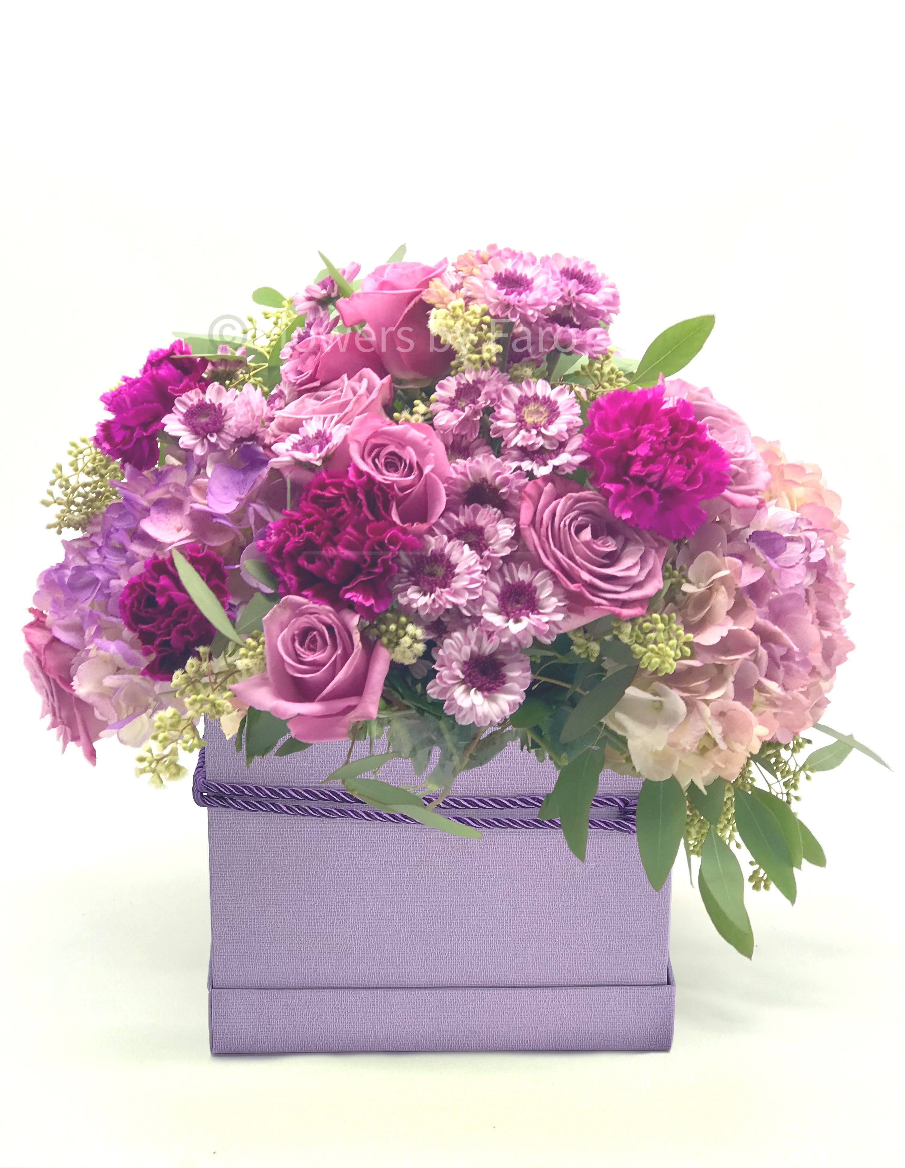 Lavender and pink roses, carnations, mums, and hydrangeas arranged in a lavender box container