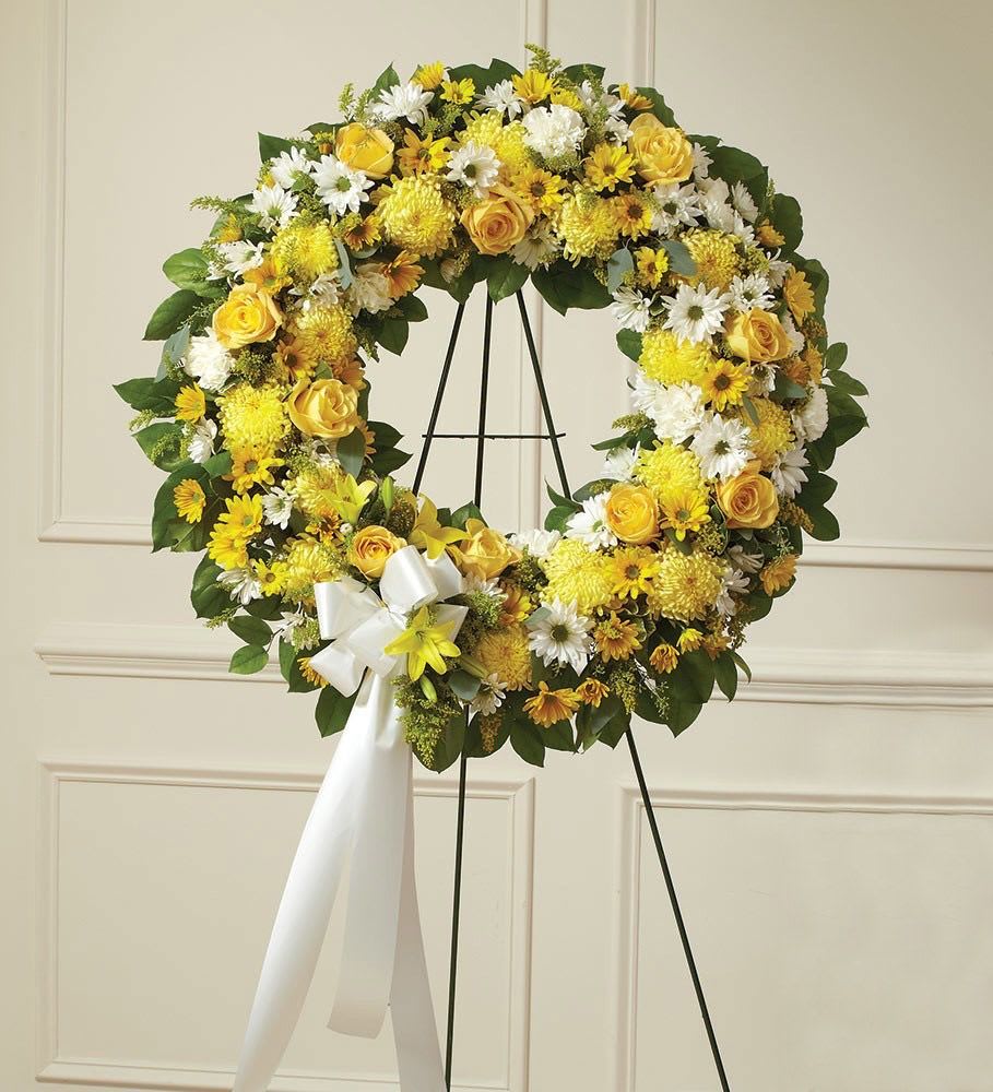 Standing wreath of yellow and white flowers with a white bow on a metal easel.