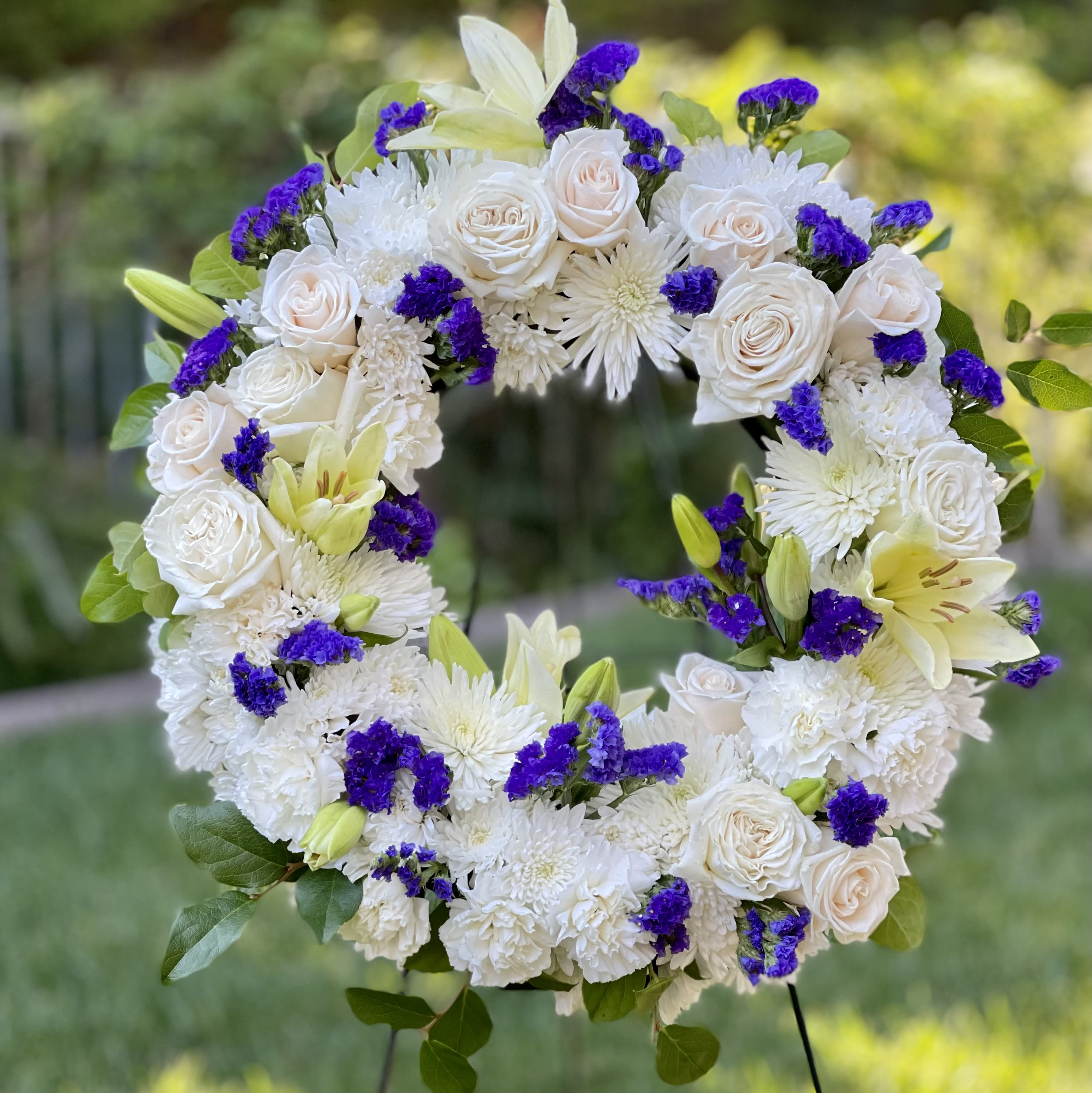 Open standing wreath of white roses, mums, and lilies with bright purple accents on a metal stand.