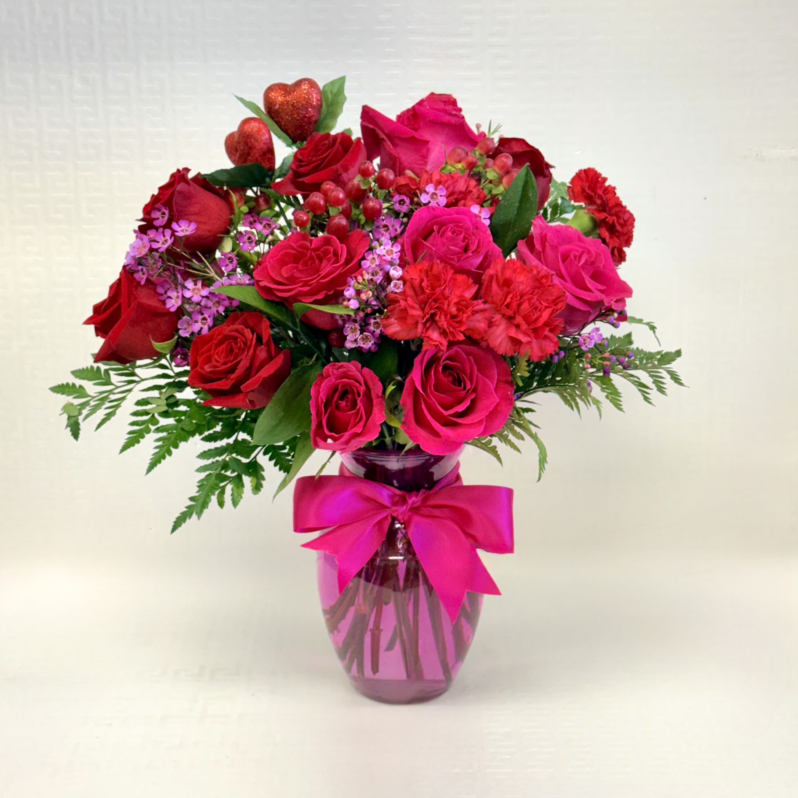 Red and hot pink roses and carnations with purple filler flowers in a pink vase with a bow.