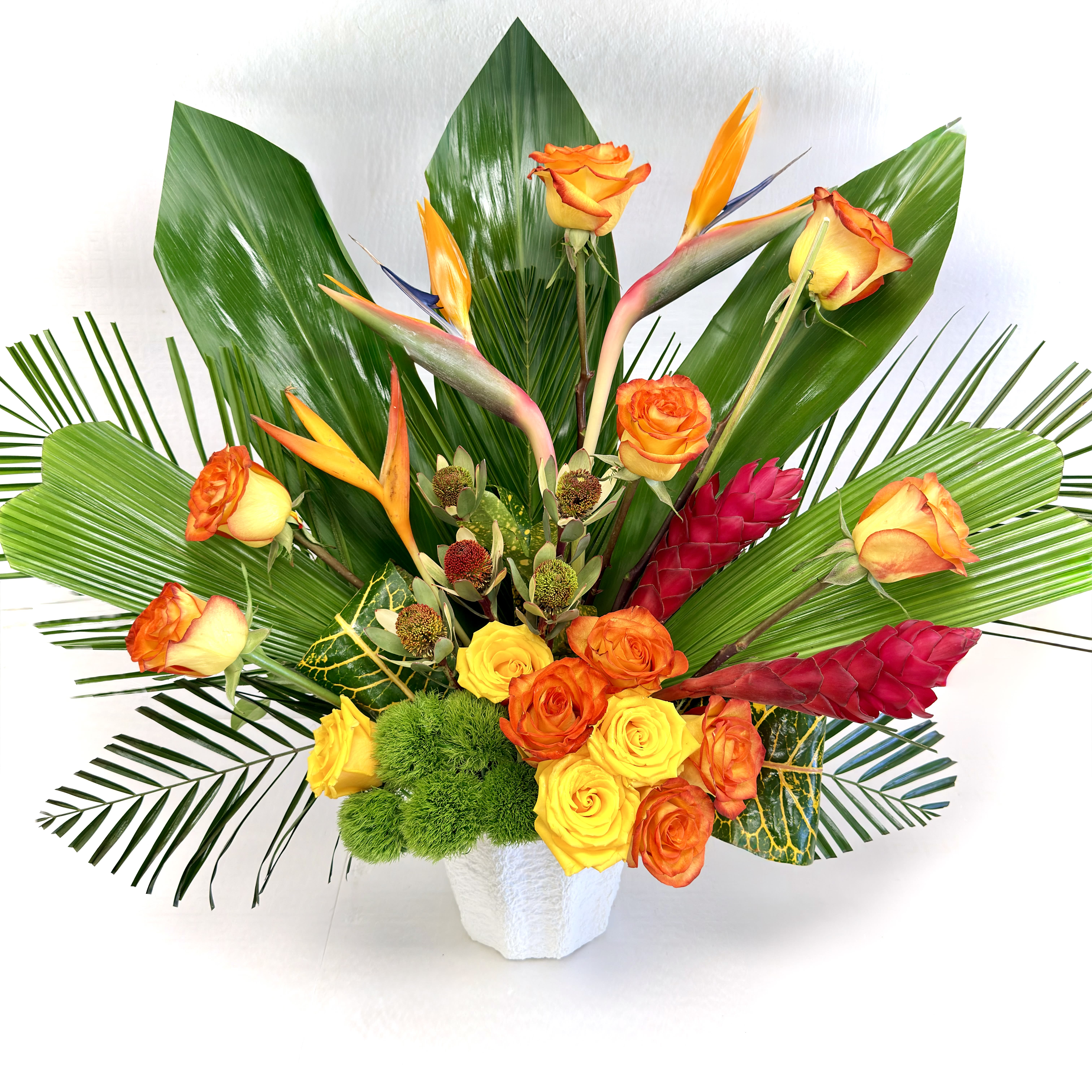 Tall tropical arrangement with orange and yellow roses and exotic blooms in a white container