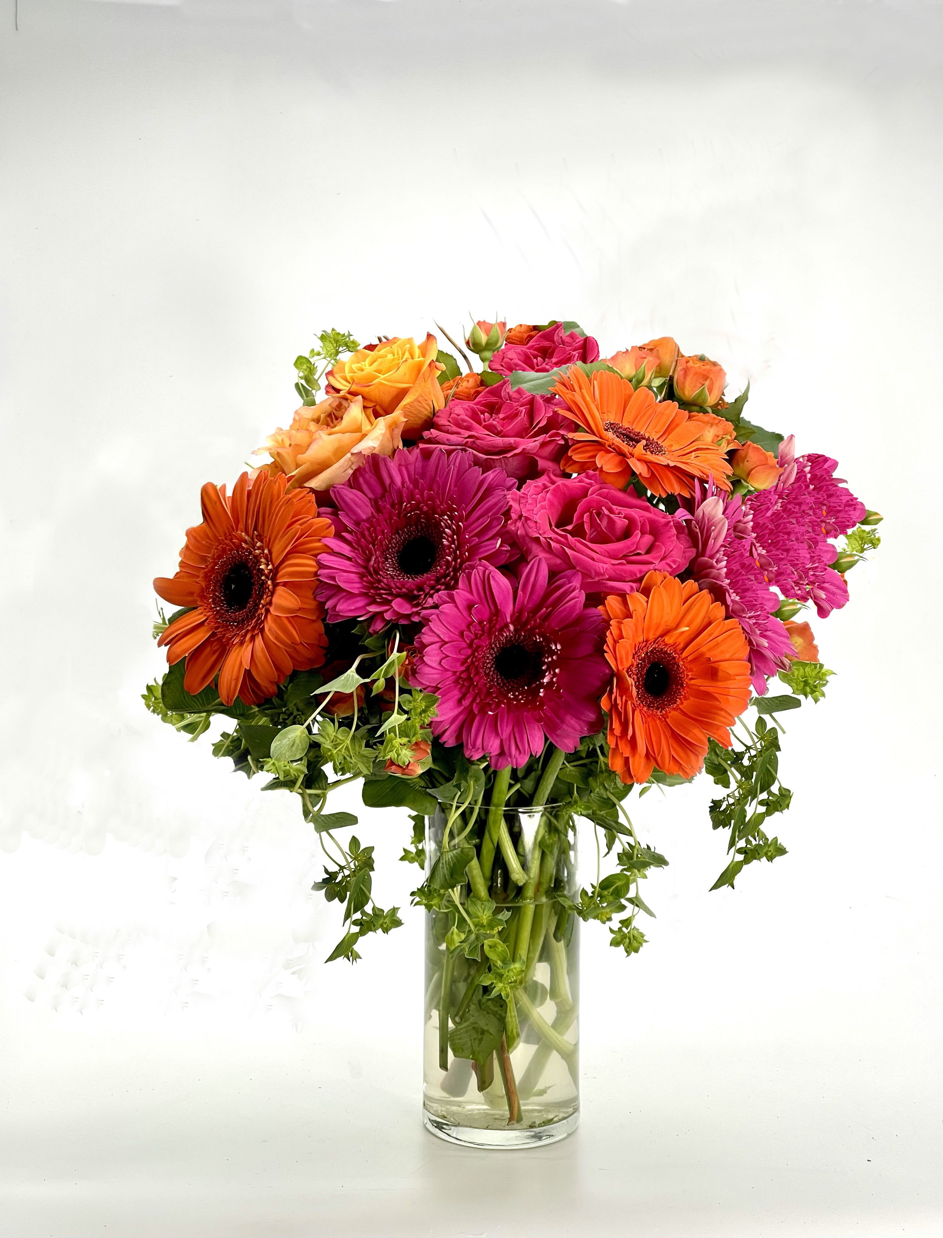 Spring Breeze Bouquet - A vibrant floral arrangement featuring bold orange gerbera daisies, bright pink roses, and lush greenery in a sleek glass vase. Perfect for adding a burst of color and elegance to any space or occasion.