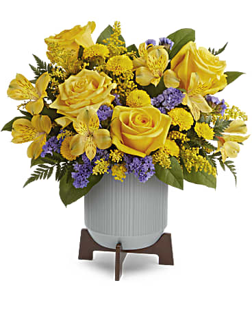 Yellow roses and yellow blooms with purple accents in a white ribbed pot on a wooden stand