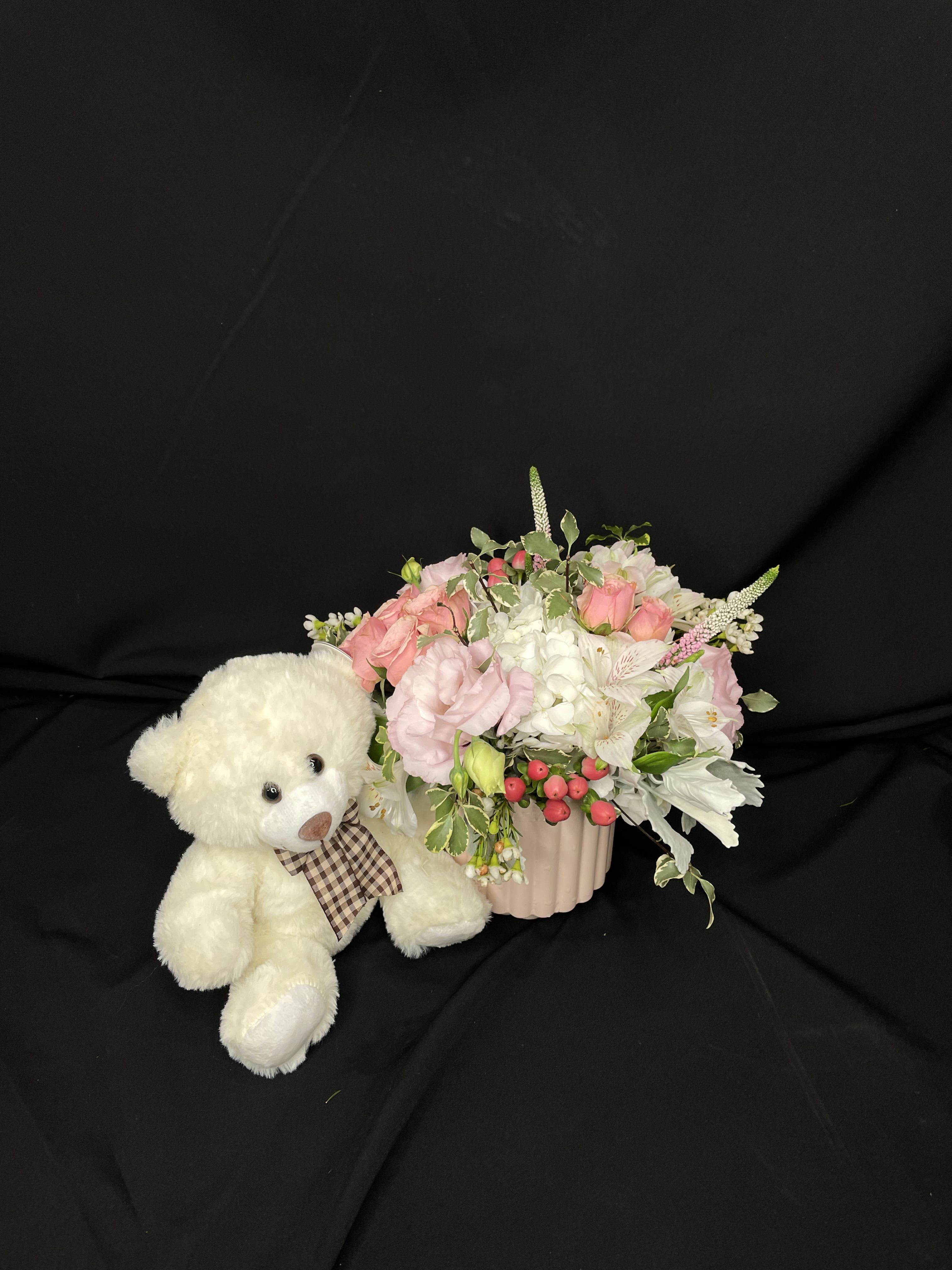 White teddy bear beside a pink floral arrangement in a ribbed vase