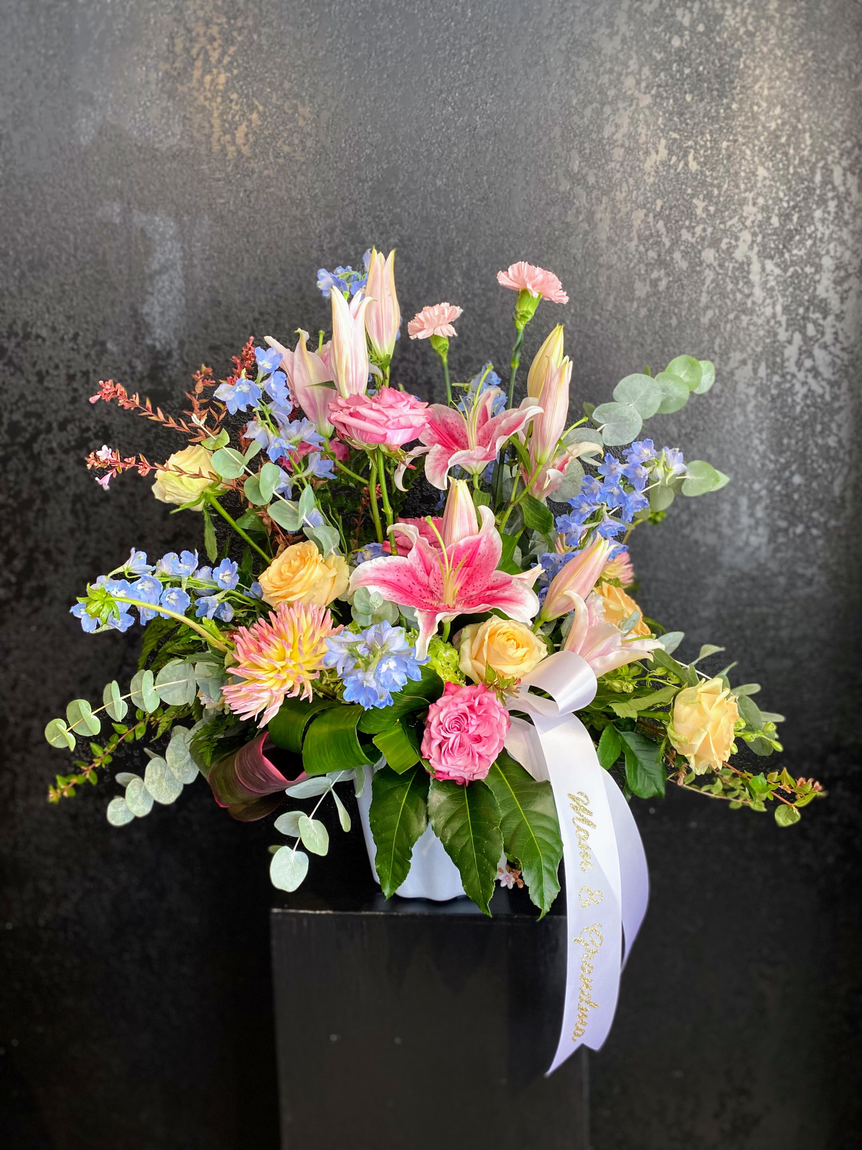 Pastel arrangement of pink lilies, roses, carnations, and blue flowers in a white vase with a white ribbon.