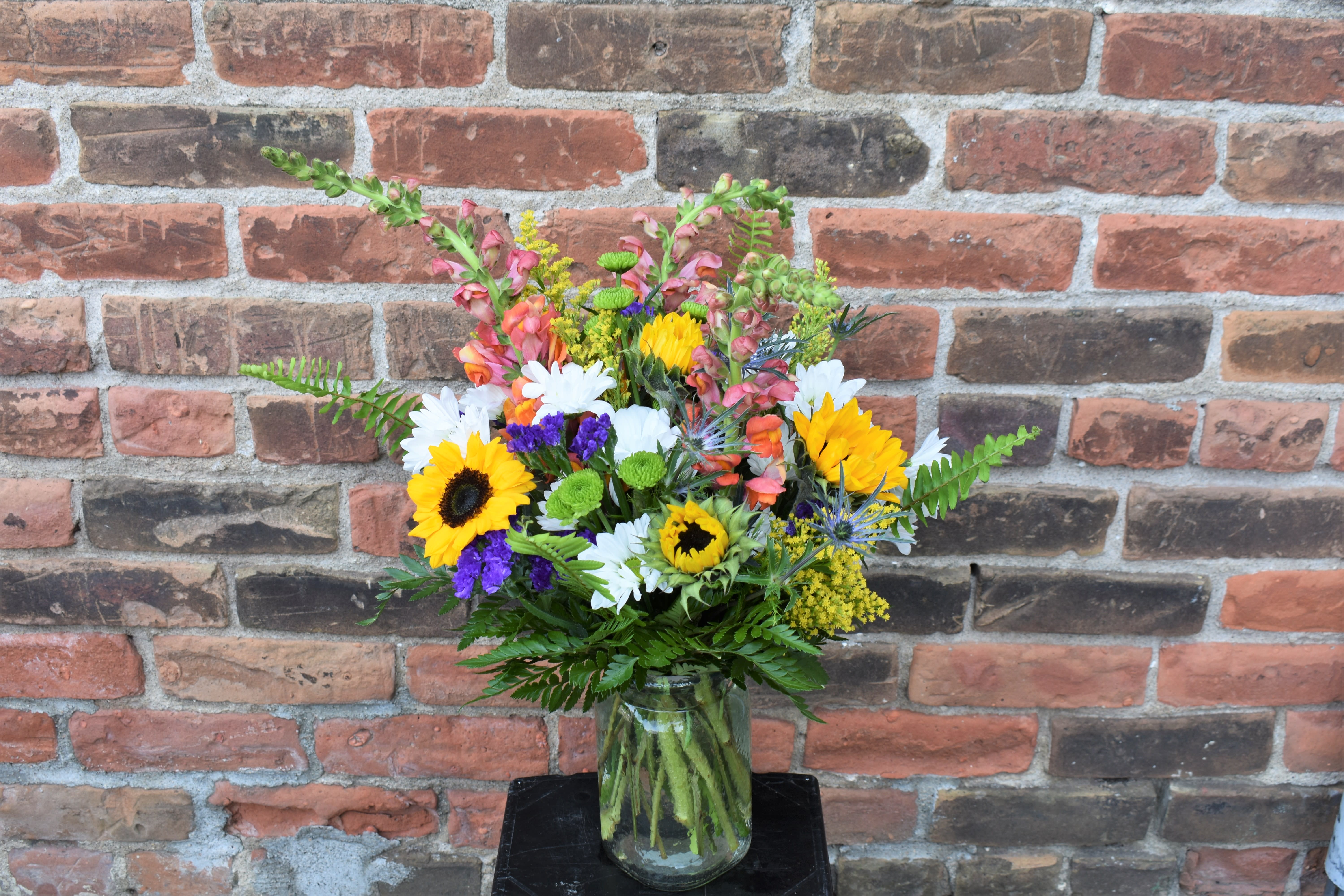 Countryside Bouquet - A bouquet bursting with an array of wildflower-themed blooms...Bright sunflowers, orange snapdragons, daisies, asters, blue thistle, and an assortment of greens are designed in a clear mason jar-styled vase...perfect for celebrating any occasion!  Size: Approx. 16"X12"
