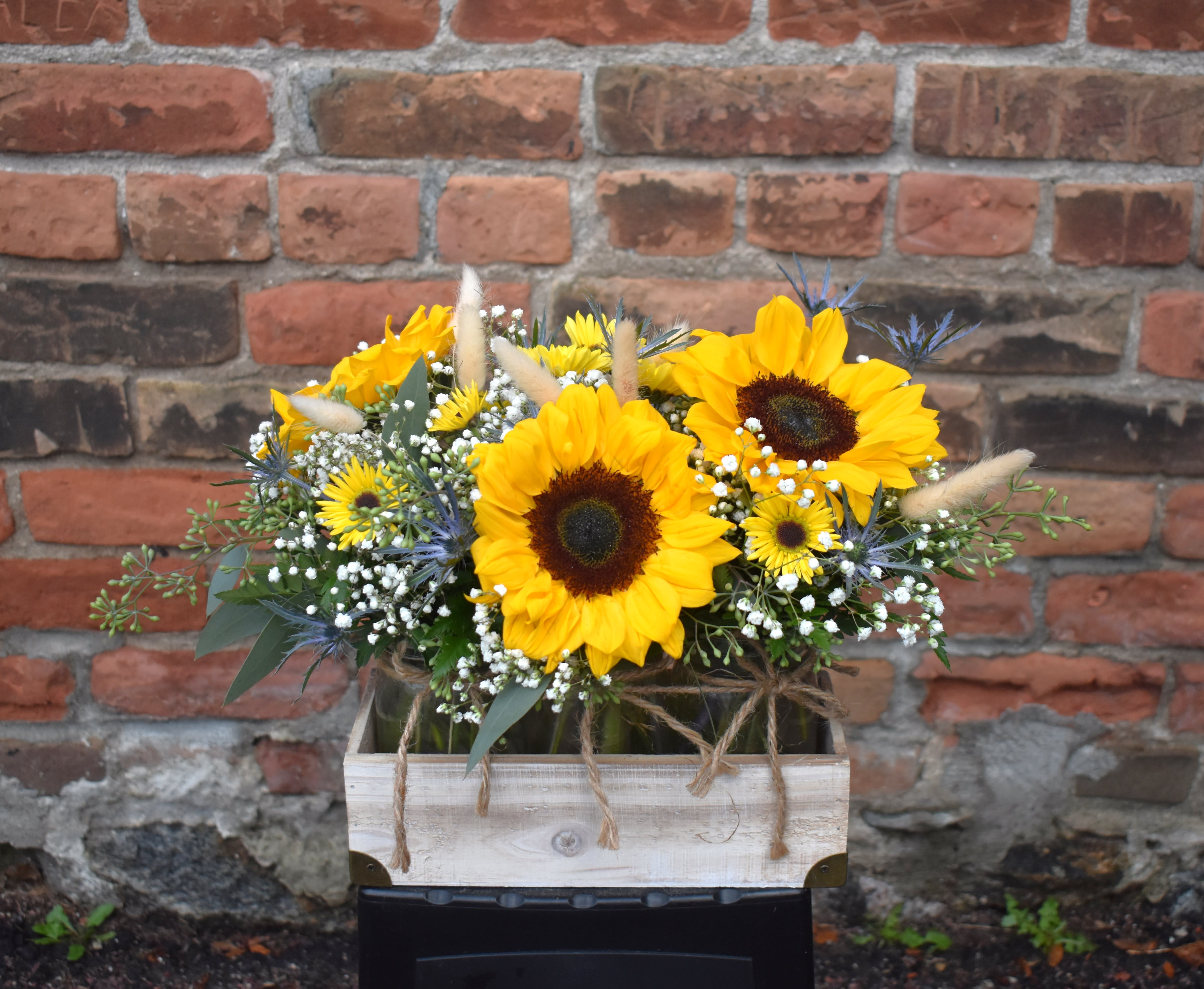 Sunshine trio - This rustic bouquet is sweet and brimming with wildflowers. Three mason jars set in a wooden box are designed with sunflowers, babysbreath, bunny tails, yellow mums, blue thistle, and eucalyptus, and accented with twine bows...the perfect way to celebrate any occasion!