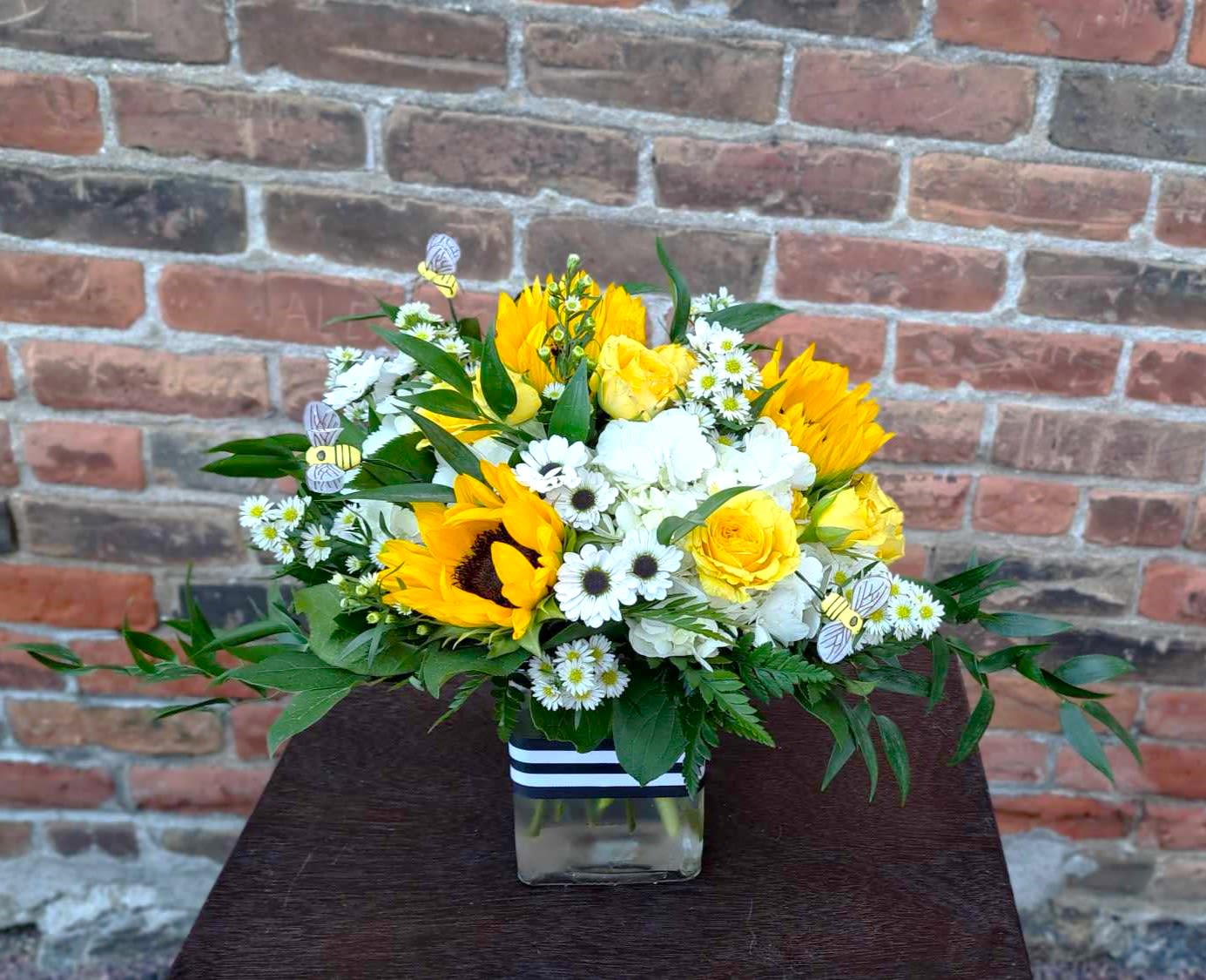 Honey Bee - This adorable bouquet is designed in a glass vase with yellow sunflowers, yellow spray roses, white hydrangea, white asters, lush greenery, and accented with cute little bees and a black/white striped ribbon band. A fun and cheery gift for any occasion! . 