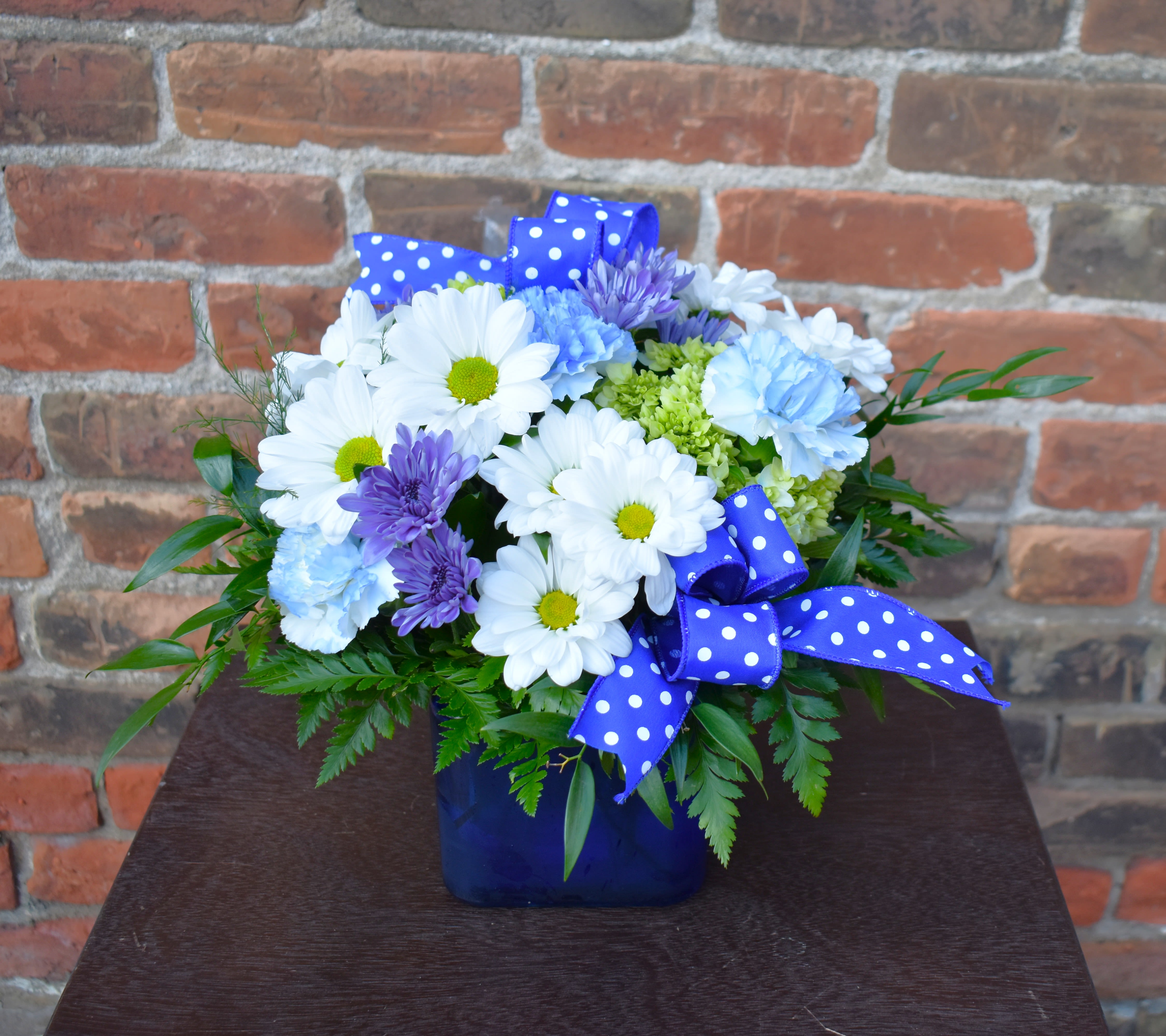 Blue vase arrangement with white daisies, purple blooms, and polka-dot ribbon