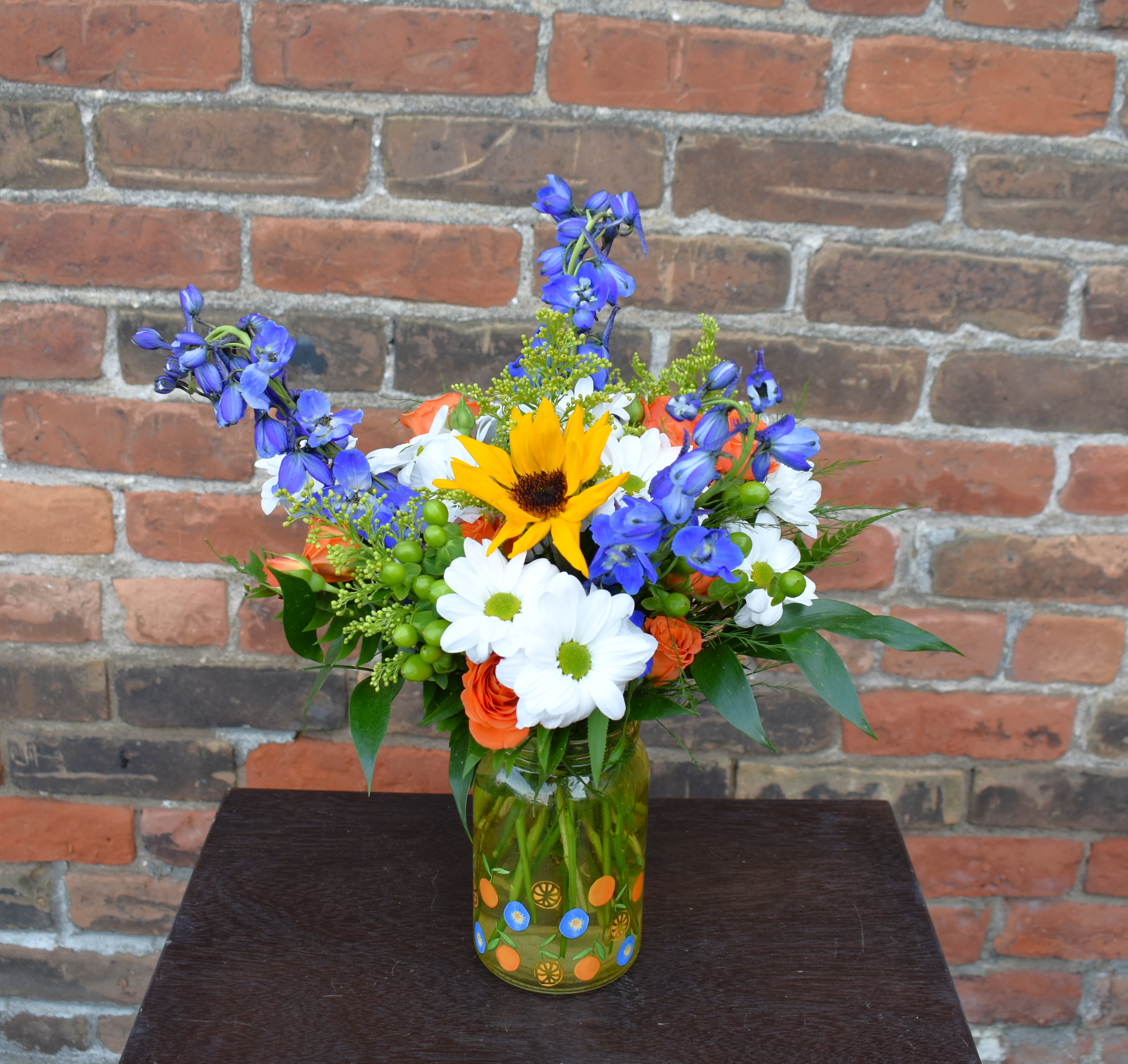 Sunshine Citrine - A summery bouquet featuring bright colors and flowers such as orange spray roses, blue delphinium, white daisies, sunflowers, green hypericum, and yellow solidago asters are designed in a vintage-style mason jar and accented with lush greens...the perfect way to celebrate any occasion! 