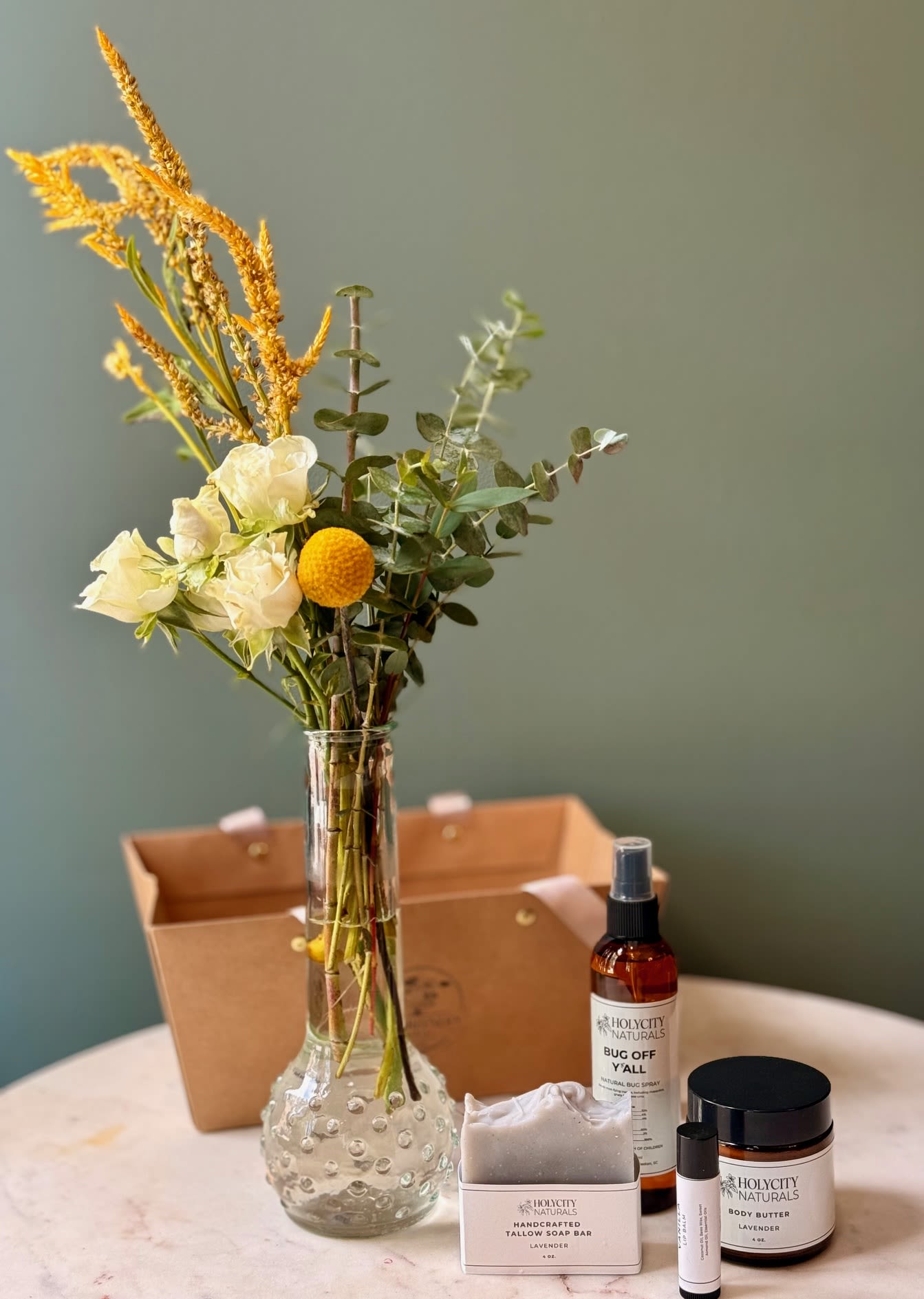 Bud Vase, Soap Bar, Body Butter, Lip Balm, &amp; Bug Spray Bundle - Designer's Choice bud vase paired with a soap bar, body butter, lip balm and bug spray in a Kraft gift basket with handles. Bud vase dimensions: approximately 10"H Products by Holy City Naturals - Hand-made in Charleston, SC.  Soap bar size: 4 Oz. Body butter size: 4 Oz. Lip Balm size: .15 Oz. Bug Spray size: 4 Oz. Available scents for soap bar and body butter: Vanilla and Lavender Lip Balm: Vanilla  * Please add your preferred scent under Special Instructions during Checkout or we will select a scent for you.  Holy City Our Naturals' collection features a lovingly handcrafted selection of soaps, bug sprays, essential oil rollers, and other natural remedies, all carefully formulated to bring more natural alternatives to your daily self-care routine.