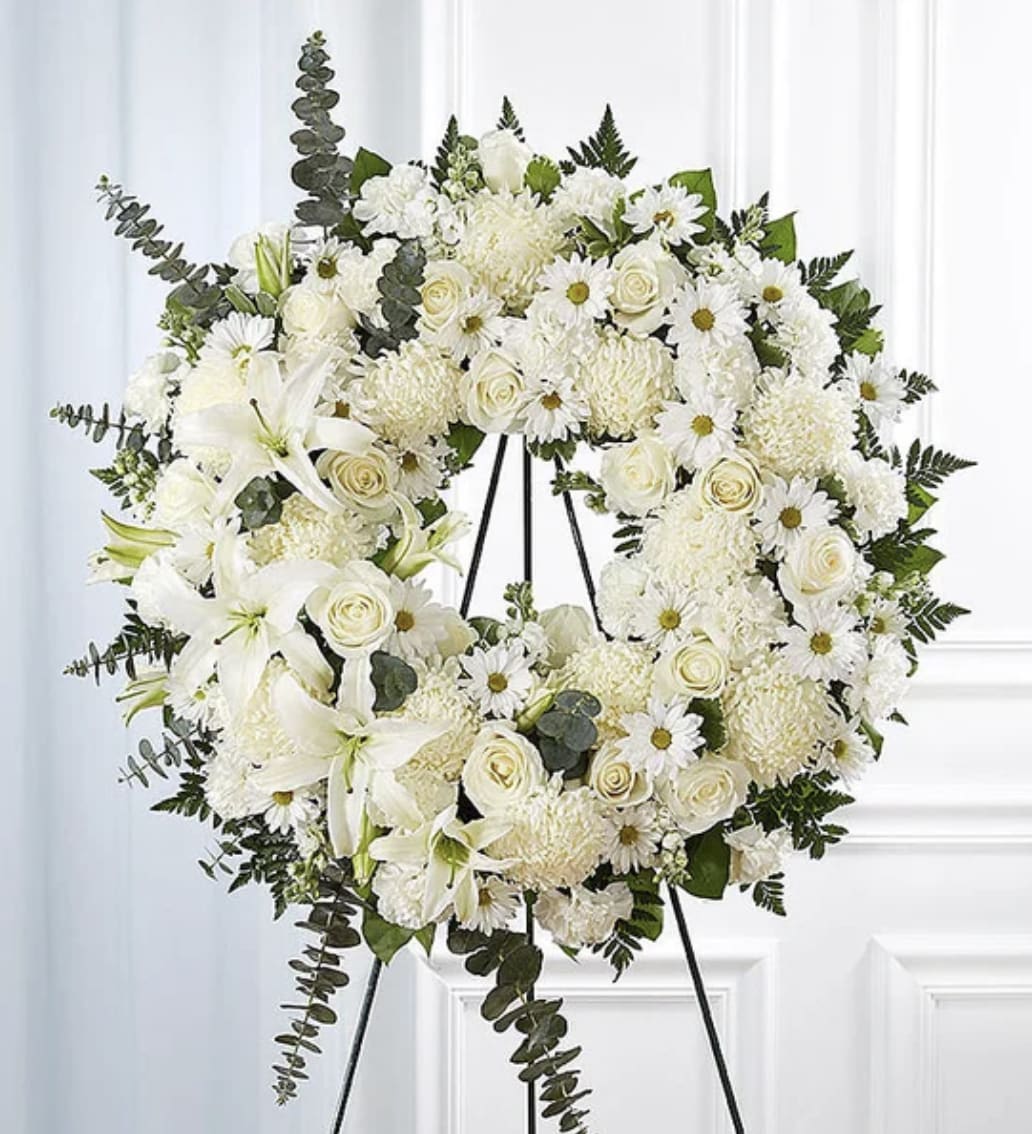 Pure Serenity Wreath - An elegant all-white wreath designed to honor with grace and timeless beauty. Featuring a lush composition of premium white blooms such as roses, lilies, carnations, chrysanthemums, and ivory seasonal blooms accented with fresh greenery, this tribute creates a serene and sophisticated display. Arranged on a standing easel, the design symbolizes peace, remembrance, purity, and everlasting love — making it a meaningful expression of sympathy and support.