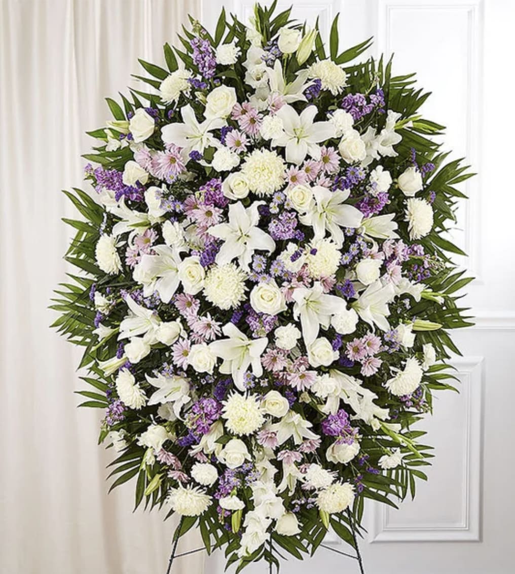 Eternal Lavender Garden Standing Spray - A graceful standing spray designed in soft white and soothing lavender tones to offer comfort, peace, and heartfelt remembrance. This elegant tribute features a lush arrangement of premium white lilies, roses, carnations and chrysanthemums, lavender daisies, mini carnations, stock, and lush seasonal blooms, accented with fresh greenery. Displayed on a standing easel, the calming color palette symbolizes serenity, grace, admiration, and everlasting love — creating a beautiful and comforting expression of sympathy.