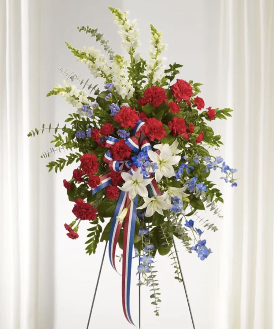 Glorious Farewell Standing Spray - A striking red, white, and blue standing spray designed to honor a cherished life with dignity, strength, and heartfelt remembrance. This patriotic tribute features an elegant arrangement of premium red roses and carnations, crisp white lilies, snapdragons and chrysanthemums, and rich blue delphinium accented with lush seasonal greenery and patriotic ribbon. Displayed on a standing easel, the bold color palette symbolizes love, peace, loyalty, and honor — creating a meaningful expression of sympathy and respect.