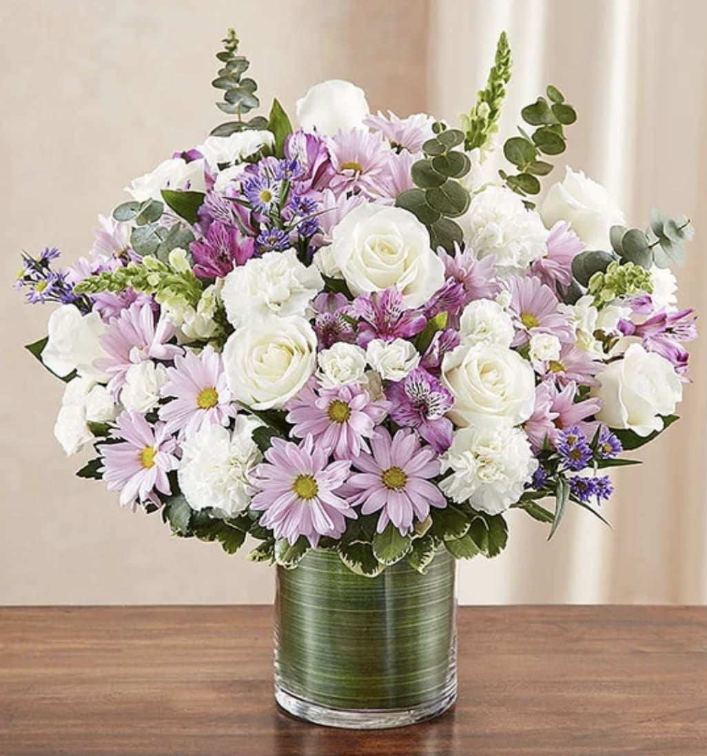 Petals of Grace - A soft and elegant bouquet designed in delicate shades of blush pink, lavender, and crisp white for a timeless and graceful presentation. This charming arrangement features lush white roses, carnations, pink daisies, alstroemeria, lavender asters, and seasonal blooms accented with fresh eucalyptus and textured greenery. Arranged in a modern leaf-lined glass vase, the bouquet offers a romantic garden-inspired feel filled with softness, beauty, and serenity. Perfect for expressing love, comfort, appreciation, or heartfelt sympathy with understated elegance.
