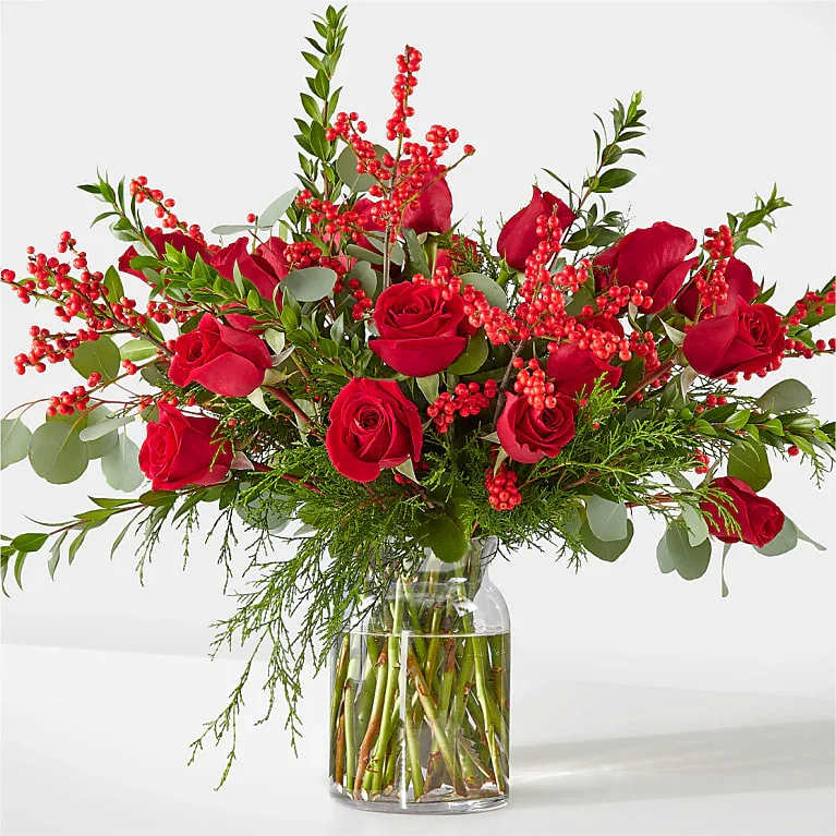 Red roses arranged in a clear glass vase with red berry branches