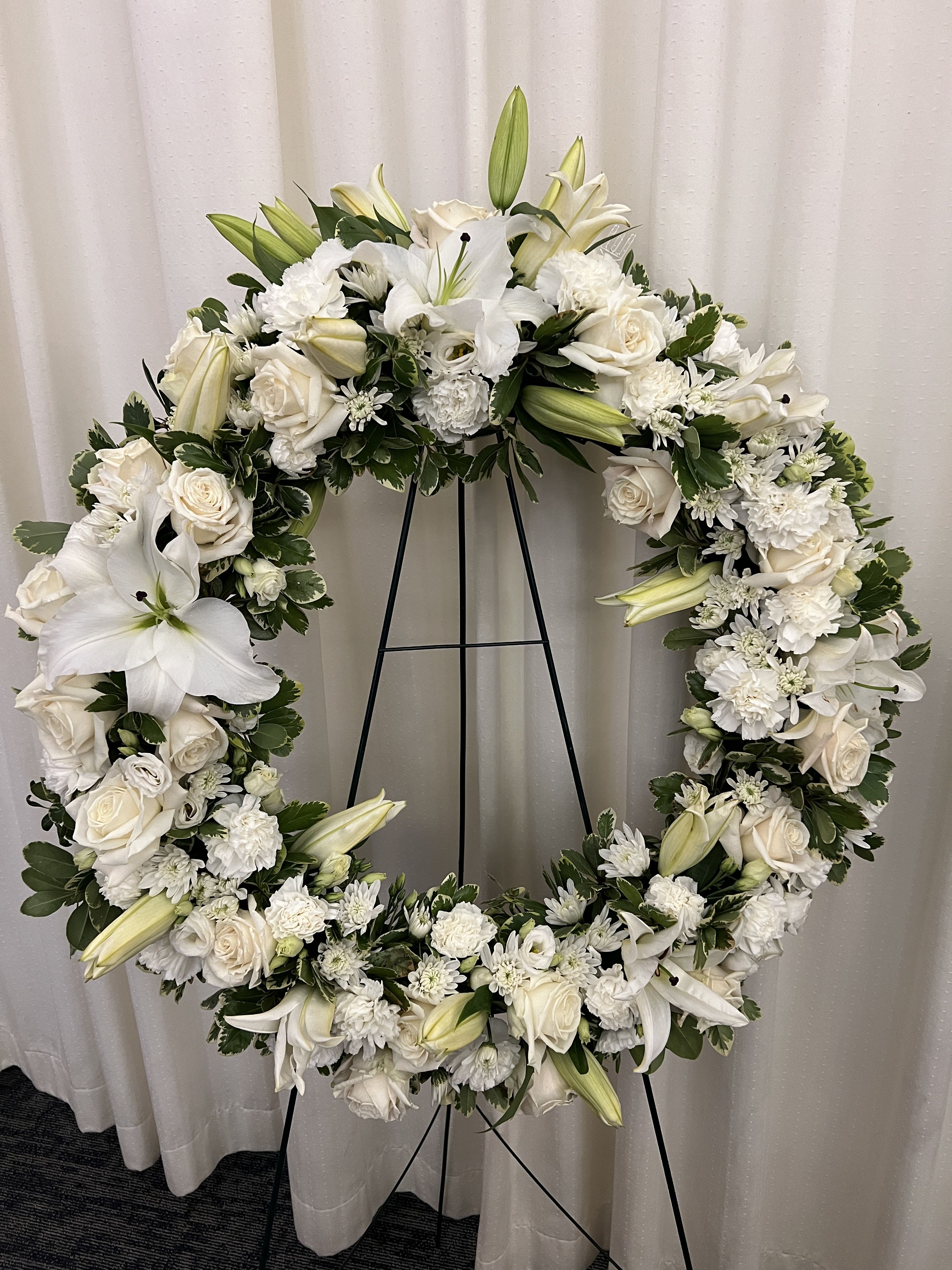White floral wreath on a black stand with roses and lilies