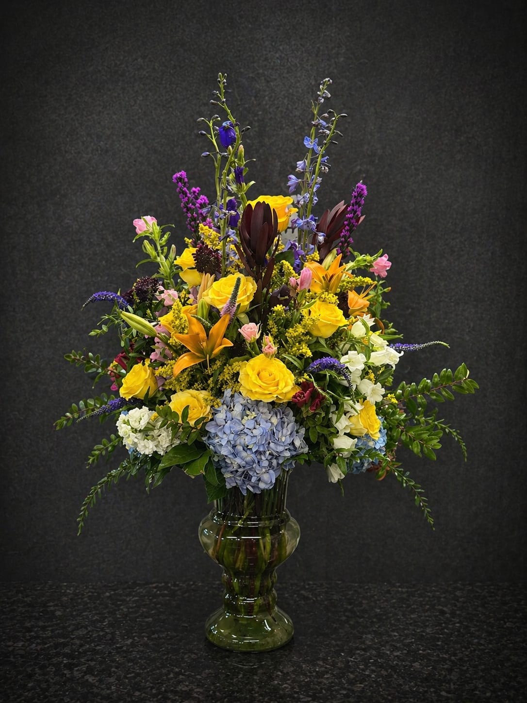 Golden Bliss - Sunlit yellow roses meet tall purple accents in a lively, garden-style bouquet that reads like a celebration in bloom-perfect as a birthday arrangement, congratulations gift, or cheerful thank-you bouquet. The upright silhouette and layered composition give the recipient an immediate sense of joy and energy; she'll feel genuinely celebrated the moment this bright, confident mix arrives.  This arrangement features bright yellow roses, pale blue hydrangea, orange lilies, white filler blooms, snapdragons and stock arranged densely in a clear glass vase. Several stems rise above the main body to create a dramatic vertical profile, while the hydrangea's soft blue tones balance the vivid yellows and purples for a fresh, textured look. Finished in a classic glass vase, it's ready to display on a mantle or entry table.  Ideal for friends, moms, coworkers, or anyone who appreciates bold color, this bouquet sends a message of warmth and upbeat encouragement. Delivered arranged and water-ready in its vase, simple daily fresh water and a cool spot will keep the blooms looking their best through the first week.
