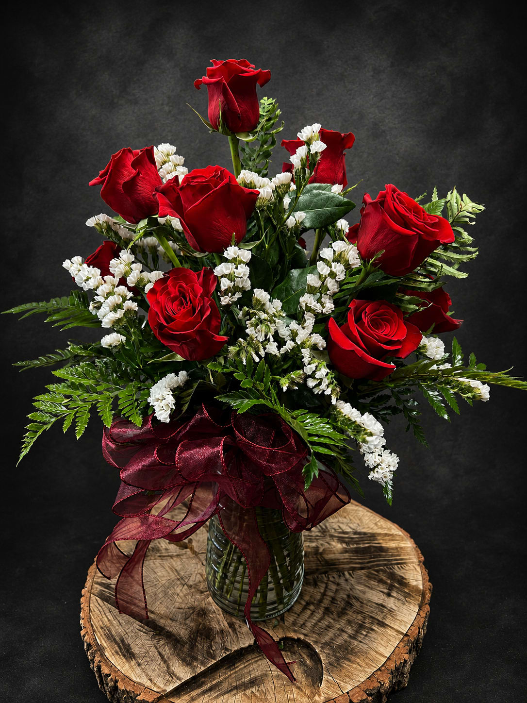 Yours Truly (dozen roses) - Deep red roses clustered with delicate white stock create a timeless dozen-rose arrangement that reads classic and romantic-perfect as an anniversary roses bouquet, a Valentine's Day surprise, or a "just because" gift to show someone they're cherished. The vivid red-and-white contrast and glossy green foliage give the person you're thinking of an immediate sense of warmth and admiration, whether you're celebrating a birthday, saying thank you, or sending love and sympathy.  Arranged upright in a clear glass vase and finished with a large burgundy ribbon, the composition is full and elegant with small white filler blooms woven through the roses for soft texture and visual lift. Expect a neatly rounded display about the size of a traditional dozen-rose design-ideal for a mantel, bedside table, or entry console. This is the kind of bouquet that makes someone feel truly seen; easy care instructions: keep water fresh, recut stems every few days, and place away from direct heat to extend bloom life.