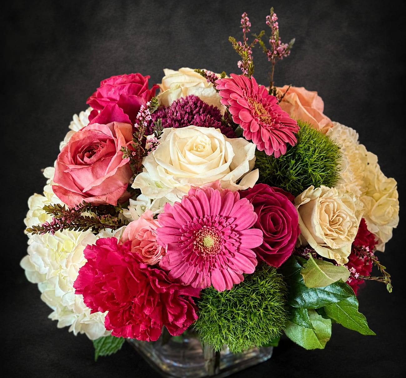 Precious Love - Soft blush and cream roses meet bright pink gerbera daisies and vivid magenta carnations in a compact, rounded bouquet designed for anniversaries, birthdays, or a heartfelt "just because" surprise. Clusters of white hydrangea and textured green dianthus give unexpected depth and contrast, while small purple filler blooms add a delicate, romantic note-the kind of arrangement that makes the person you're thinking of feel truly seen and celebrated.  Arranged tightly in a clear glass cube vase (also offered in a classic clear glass round vase), this full, cheerful design reads as both contemporary and timeless. The palette of pinks, cream, white, green and a touch of red and purple keeps it versatile for get-well wishes, housewarming gifts, thank-you gestures, Mother's Day, or a thoughtful thinking-of-you moment. Sizes are made to sit neatly on a bedside table or mantel; standard care is to change the water every two days and keep blooms out of direct heat for longest enjoyment.
