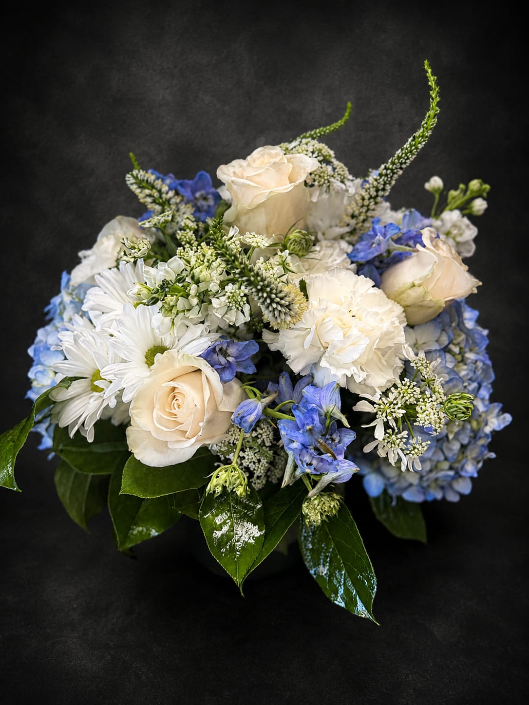 Sweet Everything  - Cream roses and airy blue hydrangeas create a calm, garden-fresh impression in this rounded floral arrangement-perfect as an anniversary bouquet or a thoughtful get well, thinking-of-you, or thank-you gift. The cool white and soft blue palette reads as both romantic and serene, so she'll feel quietly celebrated whether you're sending love, an "I'm sorry," or a cheerful housewarming surprise.  This mixed bouquet layers cream roses, white daisies, pale blue hydrangea and blue delphinium-like accents with glossy dark foliage for contrast and texture. The full, slightly rounded composition includes spray roses, stock, carnations and small blue accent flowers for a varied, natural look that feels refined without being formal. Sized to sit beautifully on a bedside table or entry console, it's a versatile choice for Spring, Admin Professionals' Week, congrats moments, or simply "just because."