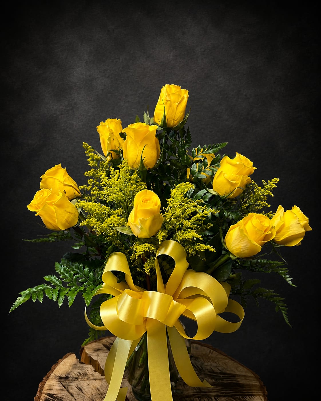 Yellow Rose of Texas - Bright clusters of yellow roses make an immediate sunny statement in this yellow rose arrangement-perfect as a birthday bouquet, anniversary gift, or cheerful get-well surprise. The rounded, upright design and the vivid yellow-on-black contrast create a polished yet joyful presence that also suits Thanksgiving centerpieces, Labor Day gatherings, or a warm "thinking of you" for Autumn and beyond.  A dozen bright yellow roses are arranged with airy yellow filler and deep green foliage in a clear glass vase, tied with a large satin yellow bow for a formal, celebratory finish. The compact, structured silhouette reads as both classic and modern, making it equally fitting for housewarmings, graduations, sympathy remembrances, or a sunny "just because" moment.  This arrangement is an easy way to make someone feel seen and celebrated-she'll feel genuinely uplifted by the cheerful color and elegant presentation. Available for same‑day local delivery when ordered early; vase and satin ribbon are included.