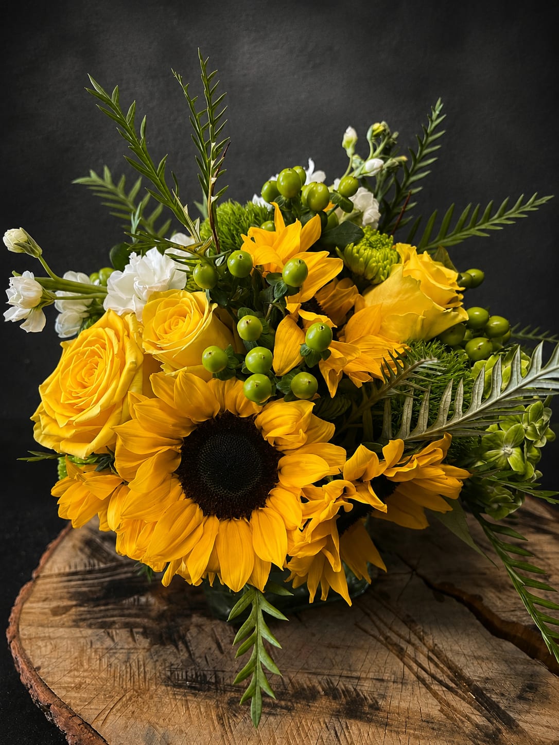 Sunshine - Golden sunflowers and sunny yellow roses take center stage in this cheerful flower arrangement for birthdays, anniversaries, graduations, or "just because" moments. The bold yellow-and-green palette-lifted by small white blooms and bright green berry-like accents-creates an immediate feeling of warmth and optimism, the kind of bouquet that makes the person you're thinking of smile the moment it arrives.  Tightly clustered blooms sit in a clear glass vase, layered for a full, garden-inspired silhouette with long, spiky foliage that adds lively texture and movement. Expect a generous, slightly rustic presentation: large sunflower heads, classic roses, stalks of stock for soft vertical interest, and hints of white to brighten the composition. The design reads as casual yet refined, perfect for thank-you gifts, housewarmings, get-well wishes, retirements, or to celebrate Mother's Day, Father's Day, or Grandparent's Day.  Made to feel fresh and easy to enjoy, this arrangement arrives ready to display; change the water every few days and trim stems for longer life. Choose this sunny bouquet when you want the recipient to feel genuinely seen-an uplifting, everyday expression for moments from congratulating a new grad to saying "I'm sorry" or "thinking of you."