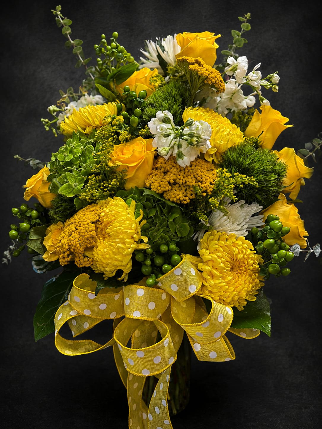 Sunset Dream - Sunlit yellow roses and chrysanthemums take center stage in this cheerful flower arrangement, a bright anniversary or birthday bouquet that instantly lifts the room. The combination of sunny yellow blooms with crisp white daisies and clustered white accents creates a joyful contrast against abundant green, textural foliage-perfect for saying "Happy Birthday," "Thank you," or "Thinking of you" with warmth. The overall effect is lively and uplifting, making it equally appropriate for spring celebrations, get well wishes, or a heartfelt just-because surprise.  This full, rounded arrangement is tied with a wide yellow polka-dot ribbon for a playful, gift-ready finish and arrives arranged in a clear glass vase for easy display. Yellow roses, yellow chrysanthemums, white daisy-like flowers and small clustered fillers mingle with berry-like and leafy greens to create depth and texture that reads fresh from every angle. Sized to fill a tabletop or entryway, it's designed to look abundant and celebratory while remaining easy to care for-fresh water and a fresh cut at delivery keep the stems happy.  Send this sunny arrangement to brighten someone's day-ideal for anniversaries, birthdays, Mother's Day, Boss's Day, or to offer comfort at a sympathy service without feeling heavy. It's the kind of bouquet that helps the person you're thinking of feel seen, cheered, and genuinely celebrated.