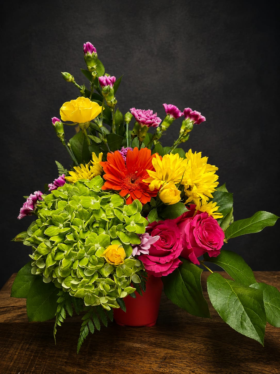 Sunshine Bright - A burst of cheerful color anchored by a red vase - Sunshine Bright is a mixed flower arrangement built to lift spirits and celebrate bright moments. This flower arrangement for Get Well, Graduation, Housewarming, or Just Because gifts combines large green hydrangea clusters with saturated pink garden roses and lively orange gerbera-style blooms so the person you're thinking of will immediately feel noticed and cheered.  The design layers texture and height: dense front-facing hydrangea and roses, with yellow daisies, chrysanthemums, pink carnations and small spray blooms rising above to create a garden-style silhouette. Colors include hot pink, sunny yellow, vivid orange and fresh green; the compact red vase gives the composition a modern, contrasted pop that reads as both festive and thoughtfully arranged.  Perfect for Spring or Summer celebrations, Thank You gestures, Thinking of You moments, Retirement congratulations, or a Romantic surprise, Sunshine Bright translates joyful intent into something tangible-she'll feel genuinely celebrated. Available in a single compact size; stems arrive arranged and ready-to-display with standard care to keep blooms fresh (change water every few days and recut stems as needed).
