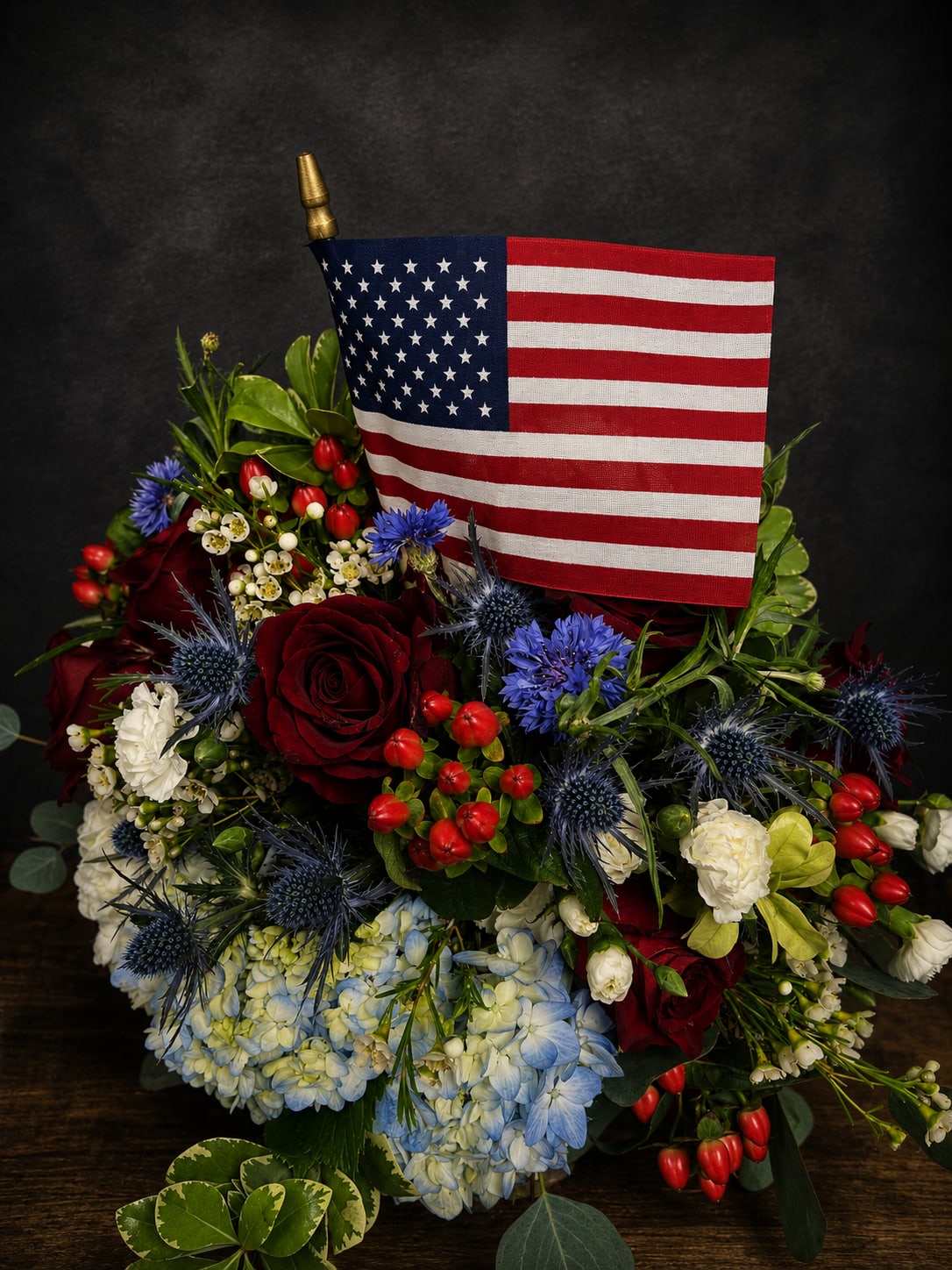 America The Beautiful - Bright, patriotic and full-bodied, this red, white and blue flower arrangement is the kind of bouquet that makes the person you're thinking of feel honored and celebrated. Designed as a housewarming or "just because" gift (and perfect for Fourth of July, Memorial Day, or retirement parties), it centers a small American flag on a compact, festive spray that reads proudly in any room.  Round, velvety red roses sit beside pale blue hydrangeas and crisp white carnations, while spiky blue thistle-like blooms and glossy red berry accents add texture and contrast. Soft stock and dianthus fill the composition for a lush, layered look; the mix of rounded petals and textured accents gives the arrangement visual depth from every angle. The flag on a short pole rises from the center for a classic patriotic touch.  Send this as a thoughtful thank-you, thinking-of-you surprise, or congratulations for promotions and retirements-the bouquet delivers clear, heartfelt intent. Sized to work on tabletops and mantels, it's ready to display on arrival; for best enjoyment, keep blooms in fresh water and away from direct heat.