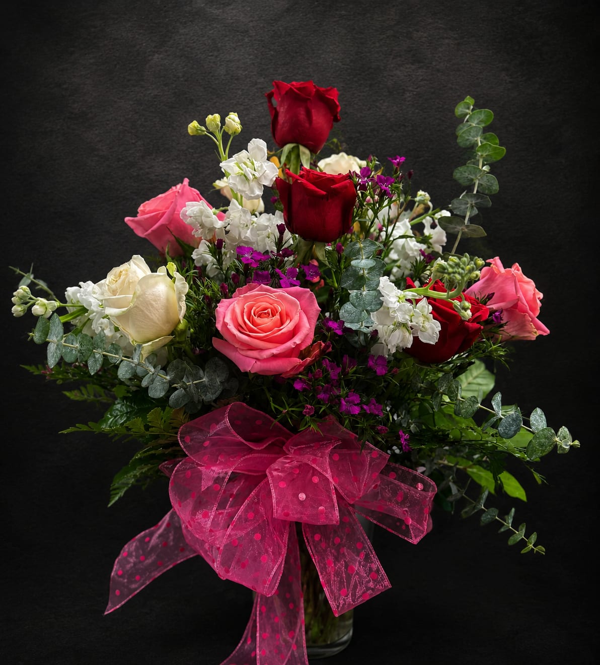 Always and Forever - A lush bouquet of red, pink and white roses creates an immediate impression of romantic celebration-perfect as an anniversary bouquet, birthday flowers, or a Valentine's Day surprise. The arrangement feels both festive and tender, making it an ideal choice for saying "I'm thinking of you" on occasions from Mother's Day and graduations to get well wishes, housewarmings, or a thoughtful sympathy gesture.  Tightly gathered in a clear glass vase, the roses are accented with small white stock-like blooms and clusters of purple filler flowers for depth and contrast. Eucalyptus-style gray-green foliage softens the palette while adding movement around the edges; a large sheer pink ribbon at the base gives the piece a polished, gift-ready finish. The composition is full and rounded with several rose heads rising above the main body for a layered, romantic silhouette.  Choose this arrangement to make the recipient feel genuinely celebrated-whether you're marking a wedding anniversary, sending thanks, offering condolences, or simply brightening someone's day just because. The clear vase and sturdy stems make care simple: fresh water regularly and a trim of stems every few days will keep the roses looking their best through spring, summer and beyond.