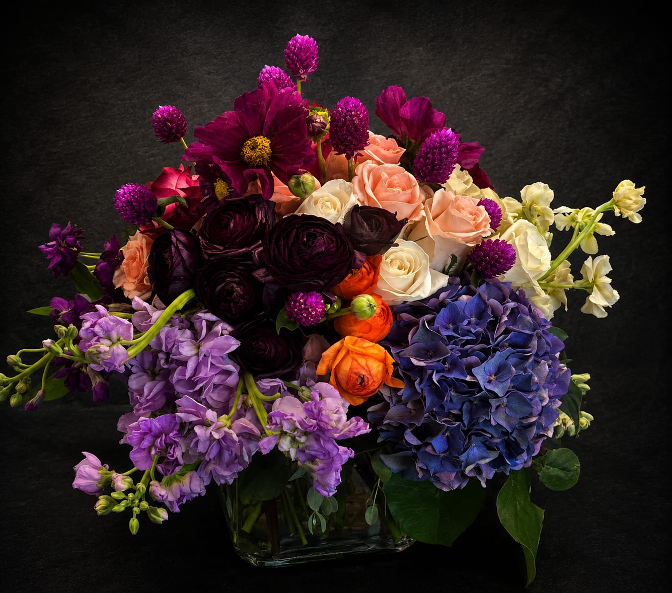 Everything to Me - Dense jewel-toned blooms in plum, lavender, blush, cream, orange and blue create an immediate sense of luxury and warmth-perfect as an anniversary bouquet, birthday surprise, or a "thinking of you" gift. This flower arrangement for special occasions like Labor Day gatherings, Thanksgiving, or a cozy autumn homecoming reads dramatic and romantic; the person you're gifting will feel truly seen by the lush, layered composition.  Large hydrangea heads anchor one side while garden-style roses and ranunculus-like blooms cluster through the center, accented by taller purple stocks that add texture and vertical interest. Presented in a clear glass vase, the compact, low-to-medium profile highlights the layered stems and lets the saturated colors pop-a bold centerpiece for holiday tables, a heartfelt get-well gesture, or an elegant housewarming display.  Ideal for celebrations from graduations and birthdays to quieter moments like sympathy, thank-you, or "I'm sorry," this arrangement works year-round and brings an everyday-luxury feeling to weddings, prom tables, or bedside bouquets for a new baby. Order by size to fit your space; change the water every two days and trim stems slightly for the longest vase life.