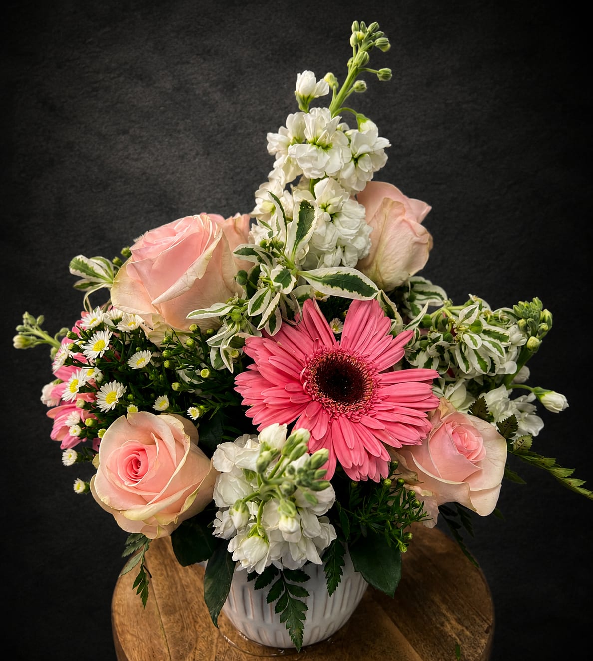 Sweetheart - Soft pink roses and a single bright pink gerbera daisy create an immediate, affectionate focal point in this sweetheart flower arrangement-perfect for an anniversary bouquet, Mother's Day gift, or a just-because surprise that says "you're cherished." The gentle pink, white, and green palette also fits spring and summer celebrations, birthdays, graduations, or a thoughtful get-well wish, and its fresh, romantic look makes it equally appropriate for sympathy and thinking-of-you moments.  The design pairs rounded rose heads with towers of white stock and small daisy-like filler blooms for a layered, garden-style texture that feels both full and airy. Arranged in a clean white ceramic vase, the composition rises in the center with smaller flowers fanning outward for balance; the compact, polished presentation fits on a bedside table, mantle, or office desk. Colors: soft pink, bright pink, white, and green; flowers: roses, gerbera daisy, stock, carnations, and daisy-like filler; vessel: white ceramic vase.  This is the kind of arrangement that makes the recipient feel truly seen-whether you're celebrating a new baby, saying thank you, marking a holiday like Labor Day or Christmas, or brightening an ordinary Tuesday. Available in a single classic size; for care, keep blooms in fresh water, trim stems every few days, and avoid direct heat to extend their life.